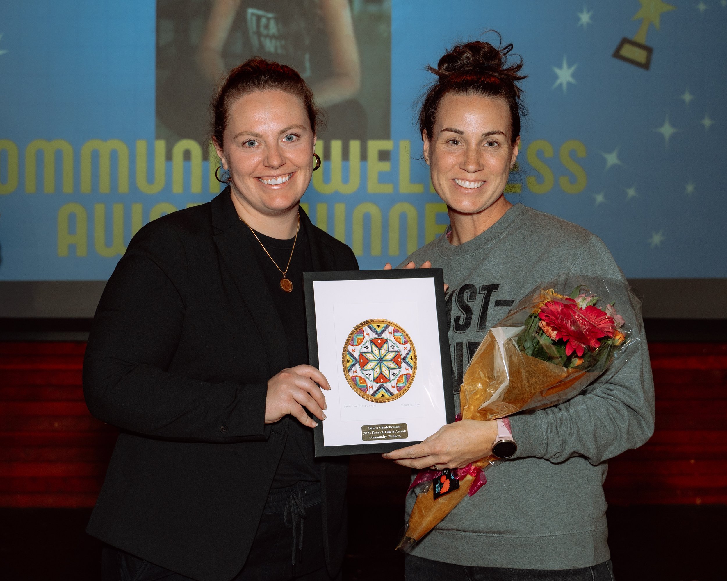 Two women smiling on stage during an award ceremony, holding a framed art piece and a bouquet of flowers, with a screen in the background displaying the words 'COMMUNITY WELLNESS AWARD'.