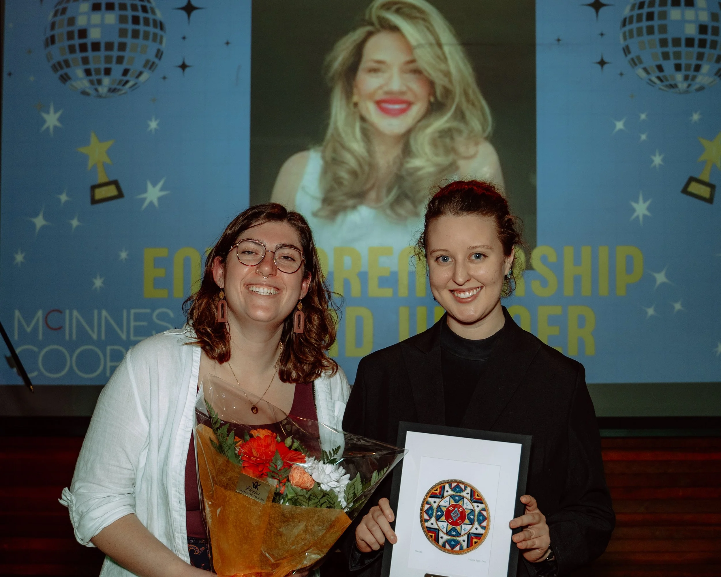 Two women smiling, one holding a bouquet of flowers and the other holding framed artwork, standing in front of a blue screen with a large projected poster featuring a woman's face and the text "EDUCATORSHIP AND HORIZON". The scene appears to be a cel