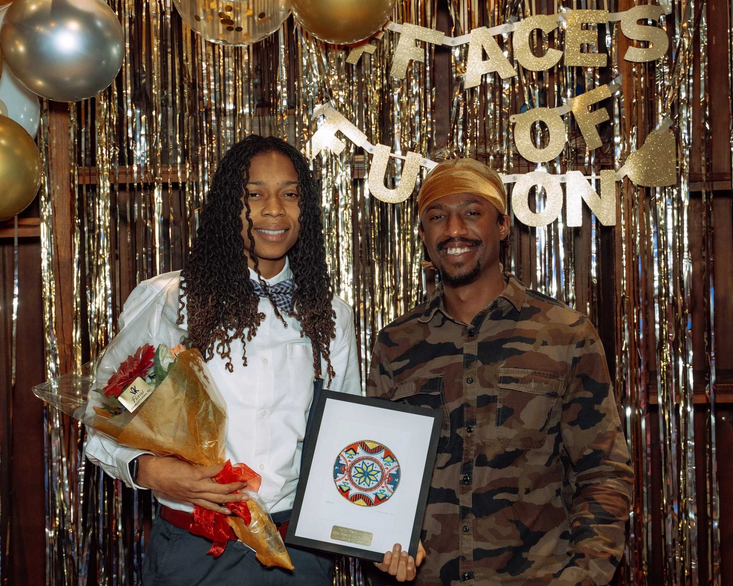 Two people standing in front of a gold and black tinsel backdrop with balloons, holding a framed artwork and a bouquet of flowers, celebrating an award or achievement at an event titled "Facets of TUON."