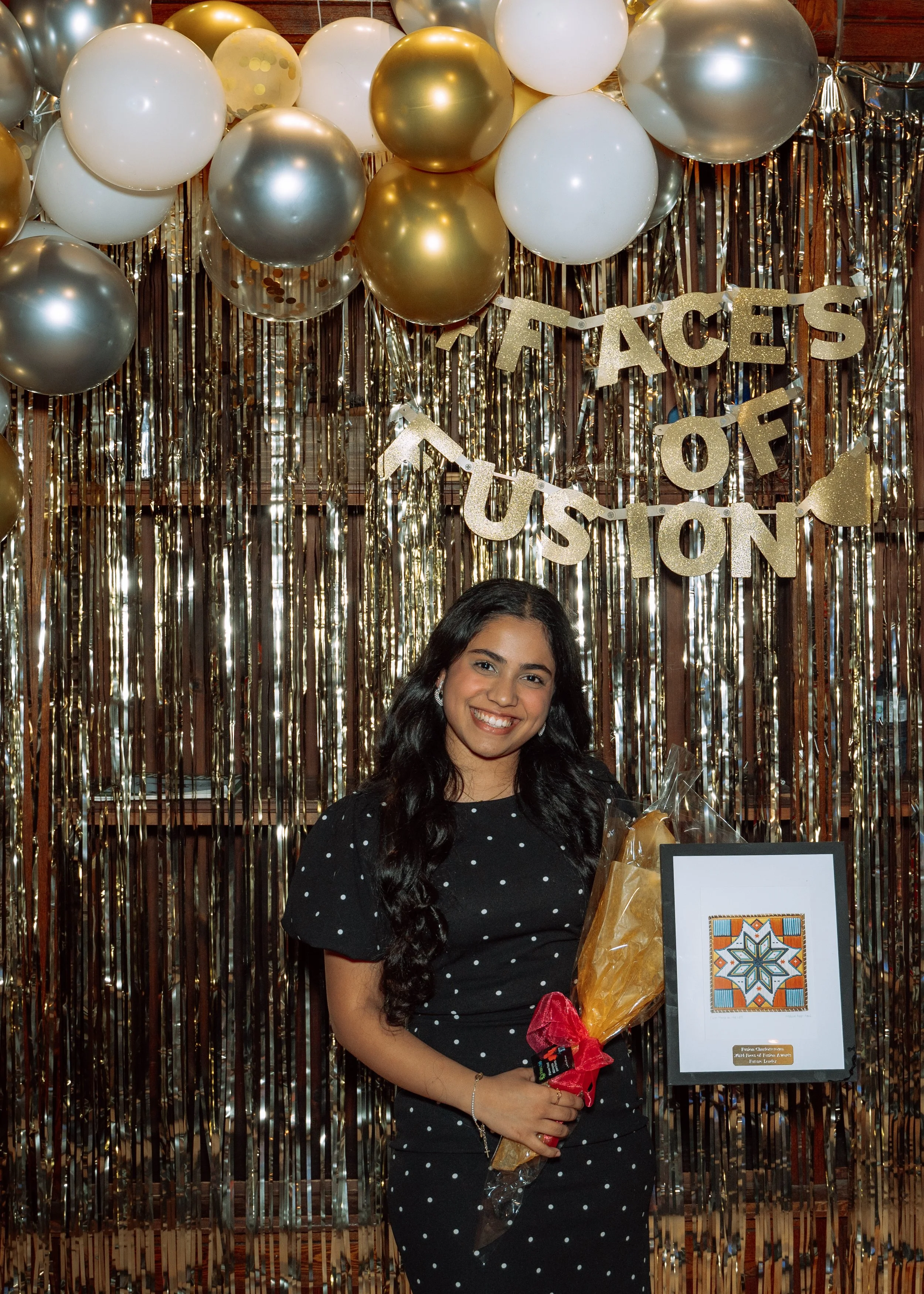 A young woman with long dark hair, wearing a black polka dot dress, is smiling and holding a wrapped bouquet of flowers. She stands in front of a festive backdrop decorated with gold, silver, and white balloons, and shiny gold and silver streamers. T