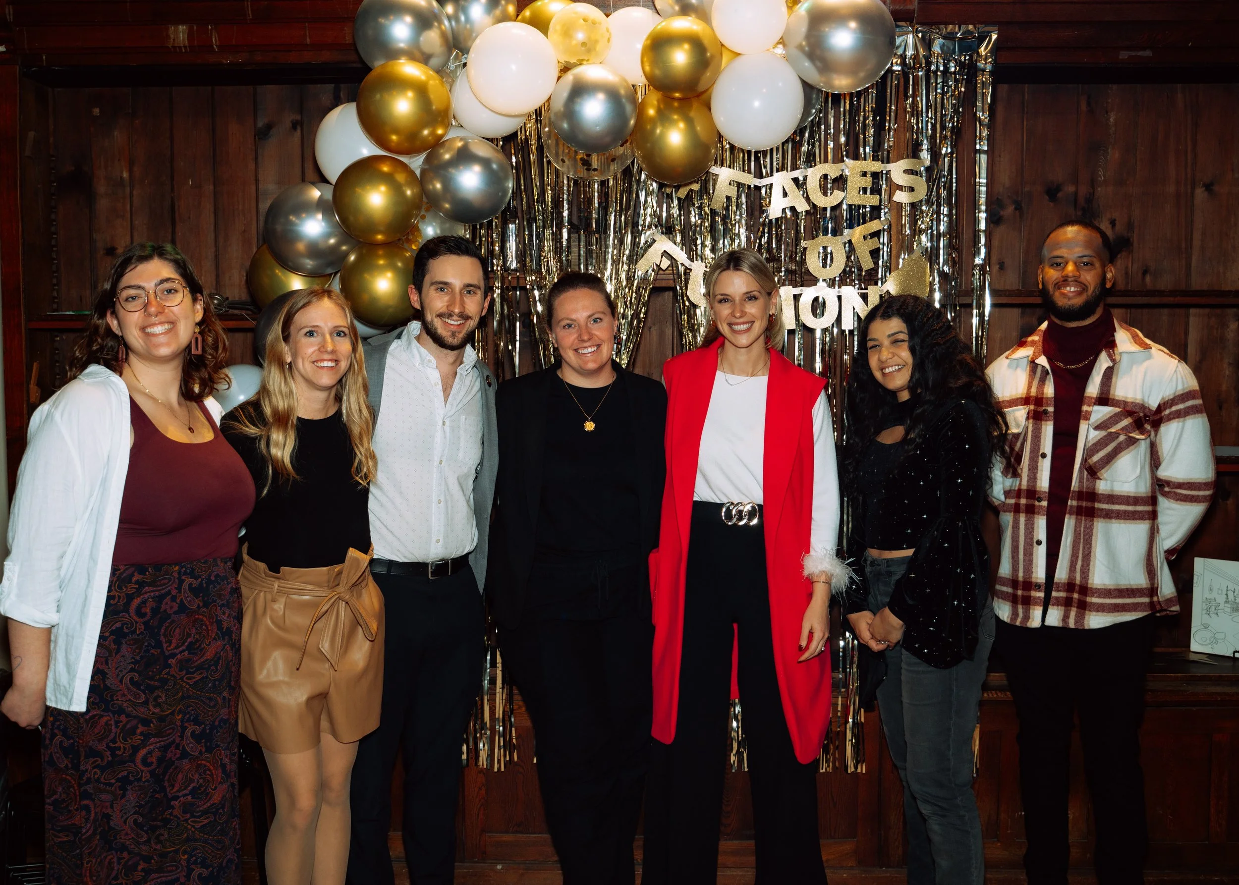 A group of seven people standing together in front of a decorative backdrop with gold, silver, and white balloons and metallic streamers. They are smiling, dressed in casual and semi-formal attire, celebrating an event with the sign 'Faces of Action'