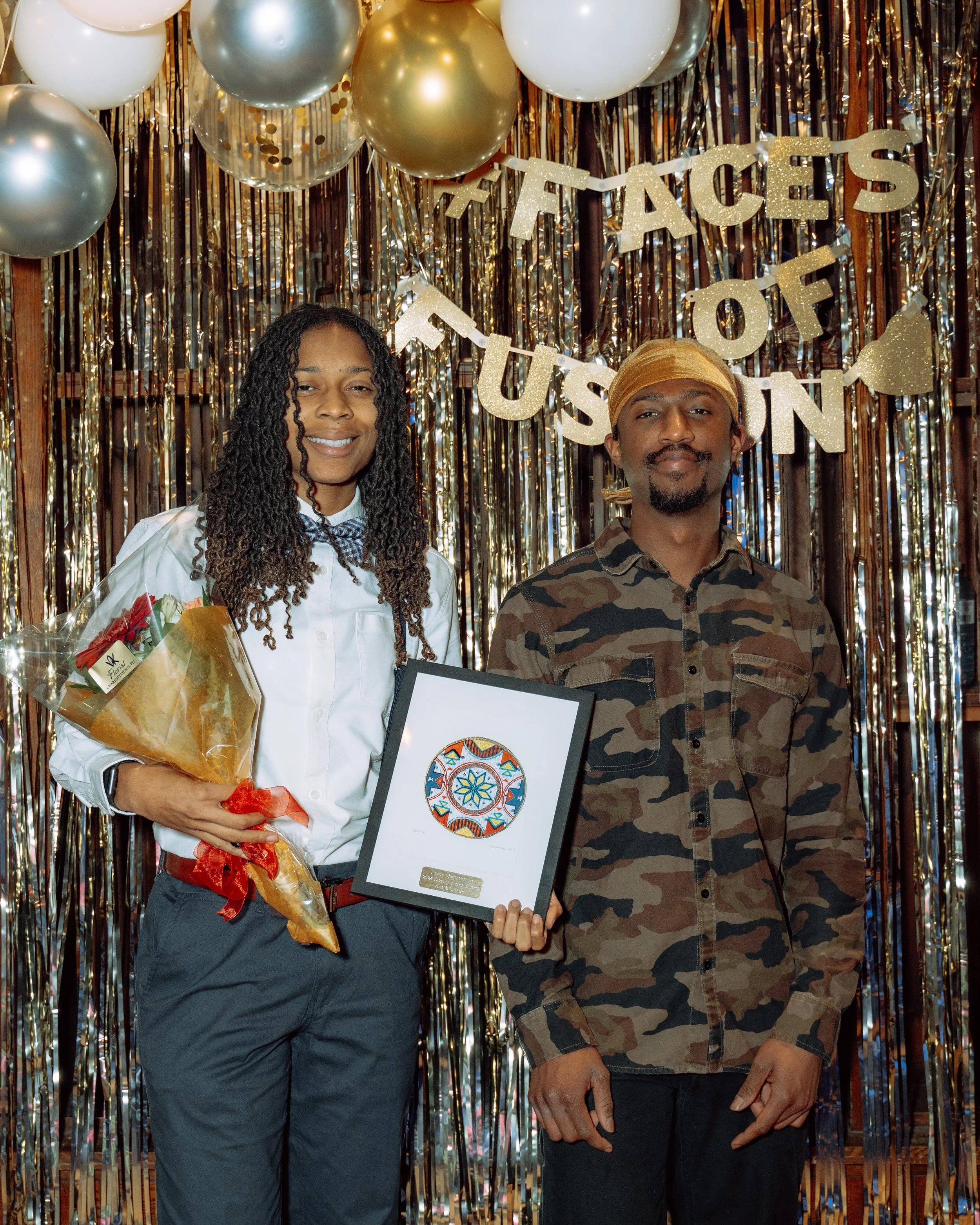 Two people standing in front of a gold tinsel backdrop with balloons and a banner that says 'Faces of Fusion.' One person is holding a bouquet of flowers and a framed certificate.
