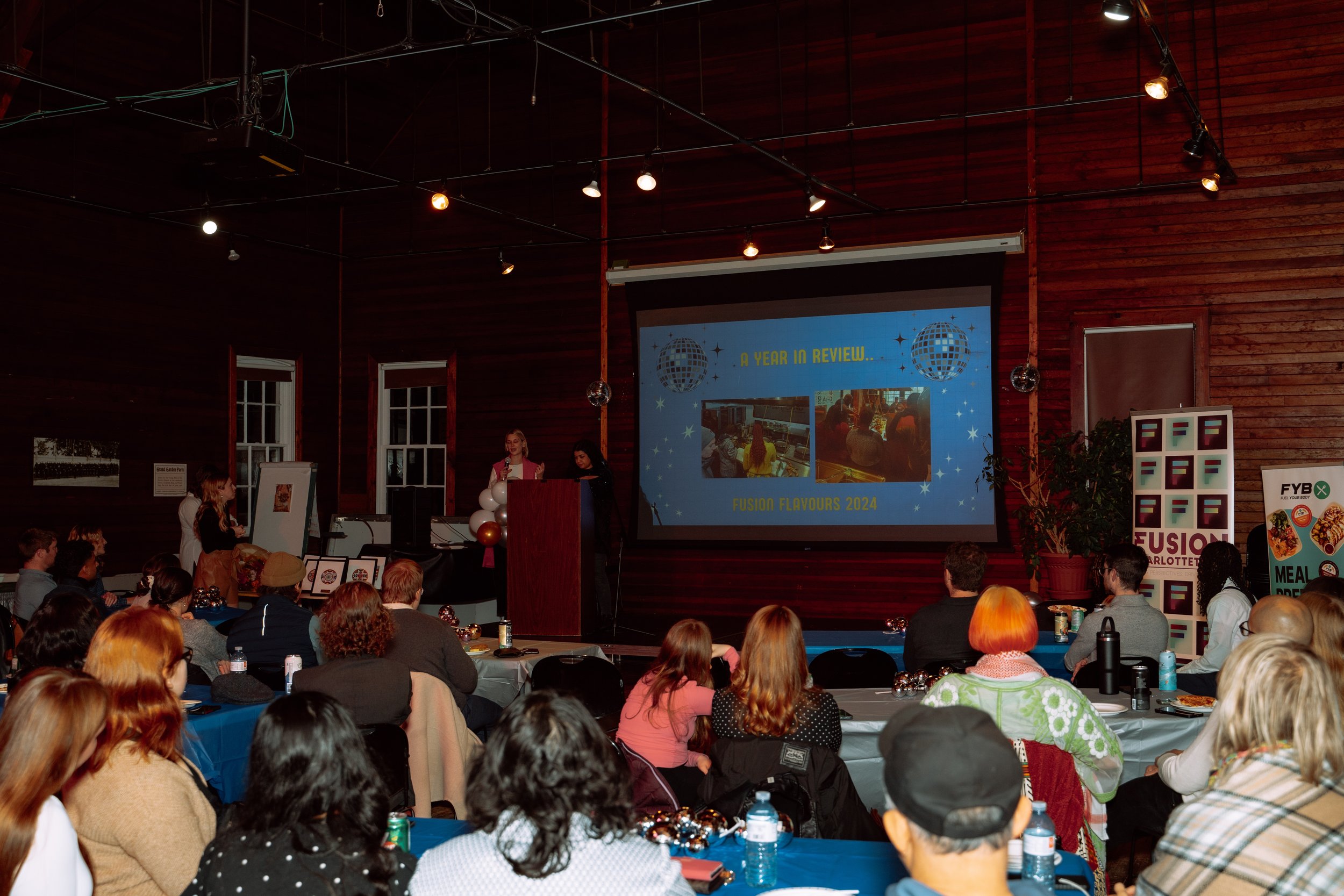 People attending a presentation in a wooden hall with a large screen displaying 'A Year in Review... Fusion Flavours 2024' and images of various food scenes.
