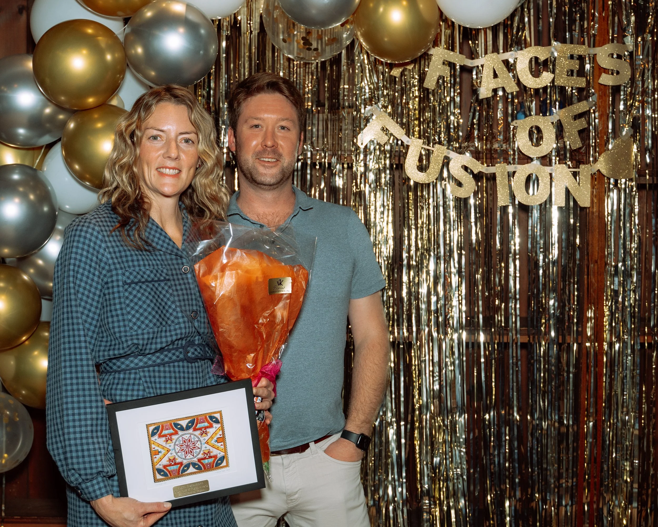 A woman and a man standing in front of a festive backdrop with gold balloons and a shiny gold fringe curtain. The woman is holding a framed colorful artwork and a bouquet of orange flowers. The backdrop has a banner that reads "Faces of Houston."