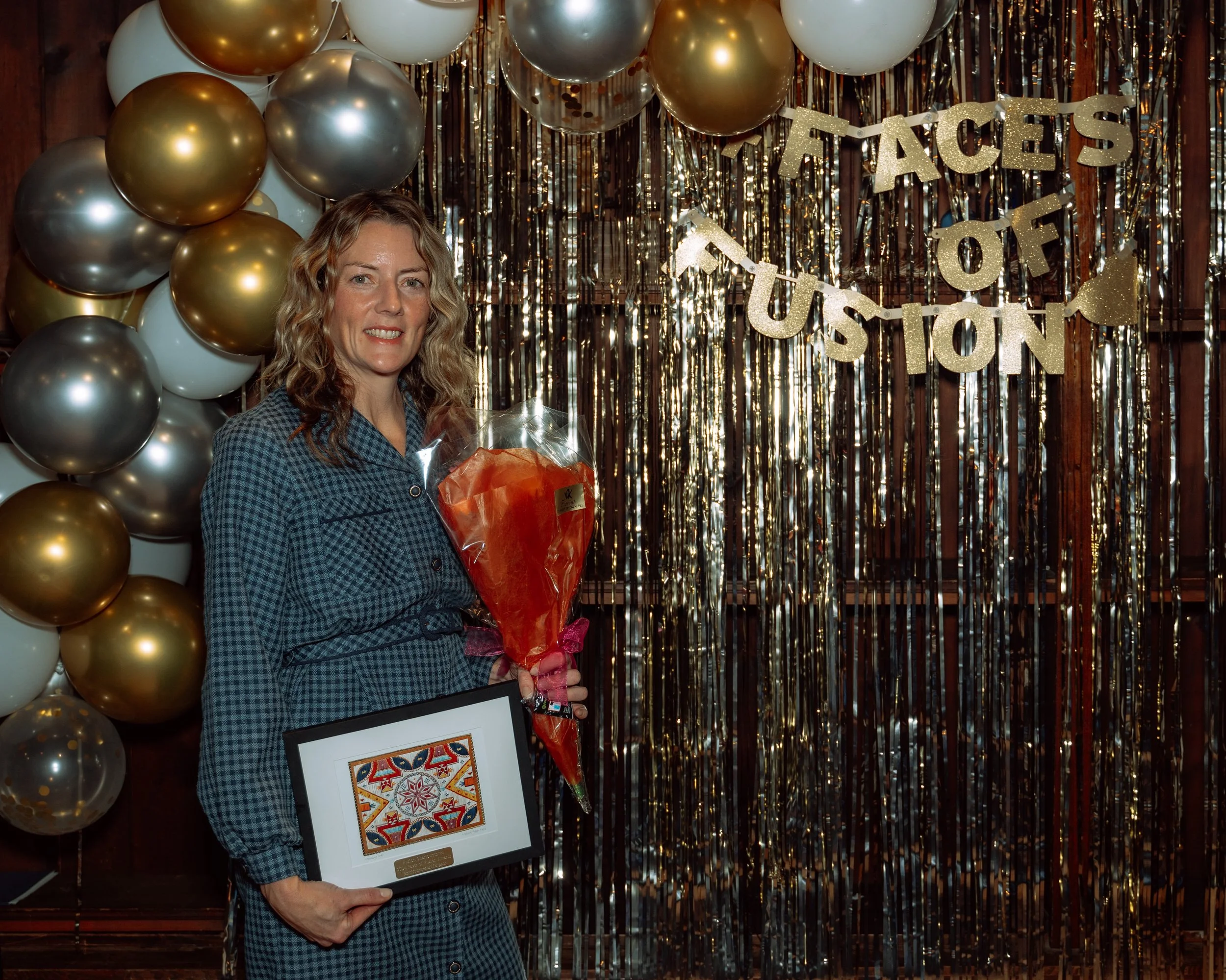 A woman in a checkered dress holding a framed colorful artwork and a bouquet of flowers, standing in front of a backdrop decorated with gold, silver, and white balloons, and a gold banner that reads 'Faces of Fusion' at an event.