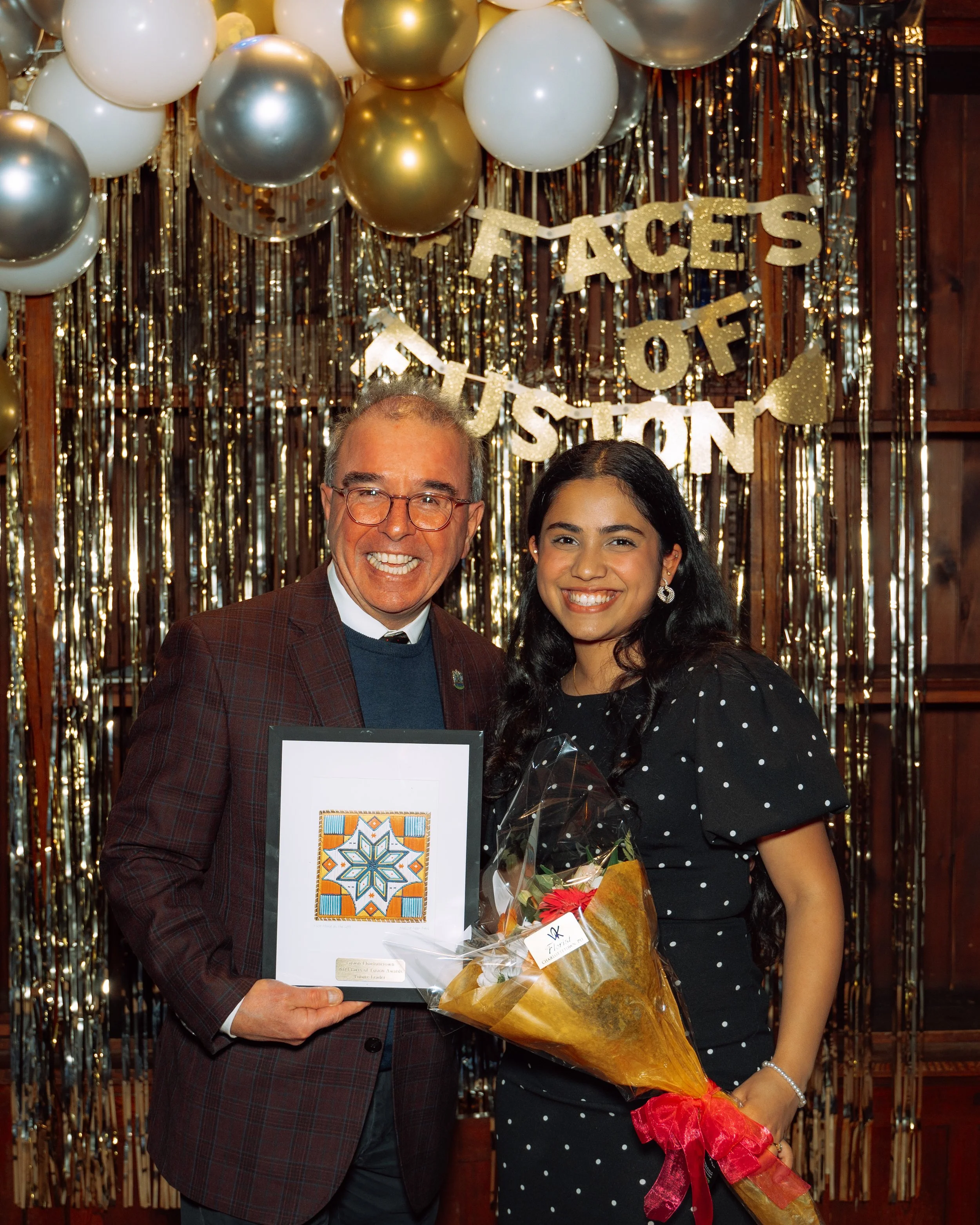 A man and a woman smiling and standing together at an event, the man is holding a framed artwork and the woman is holding a bouquet of flowers. Behind them is a backdrop with balloons and a decoration that reads "Faces of Fashion".