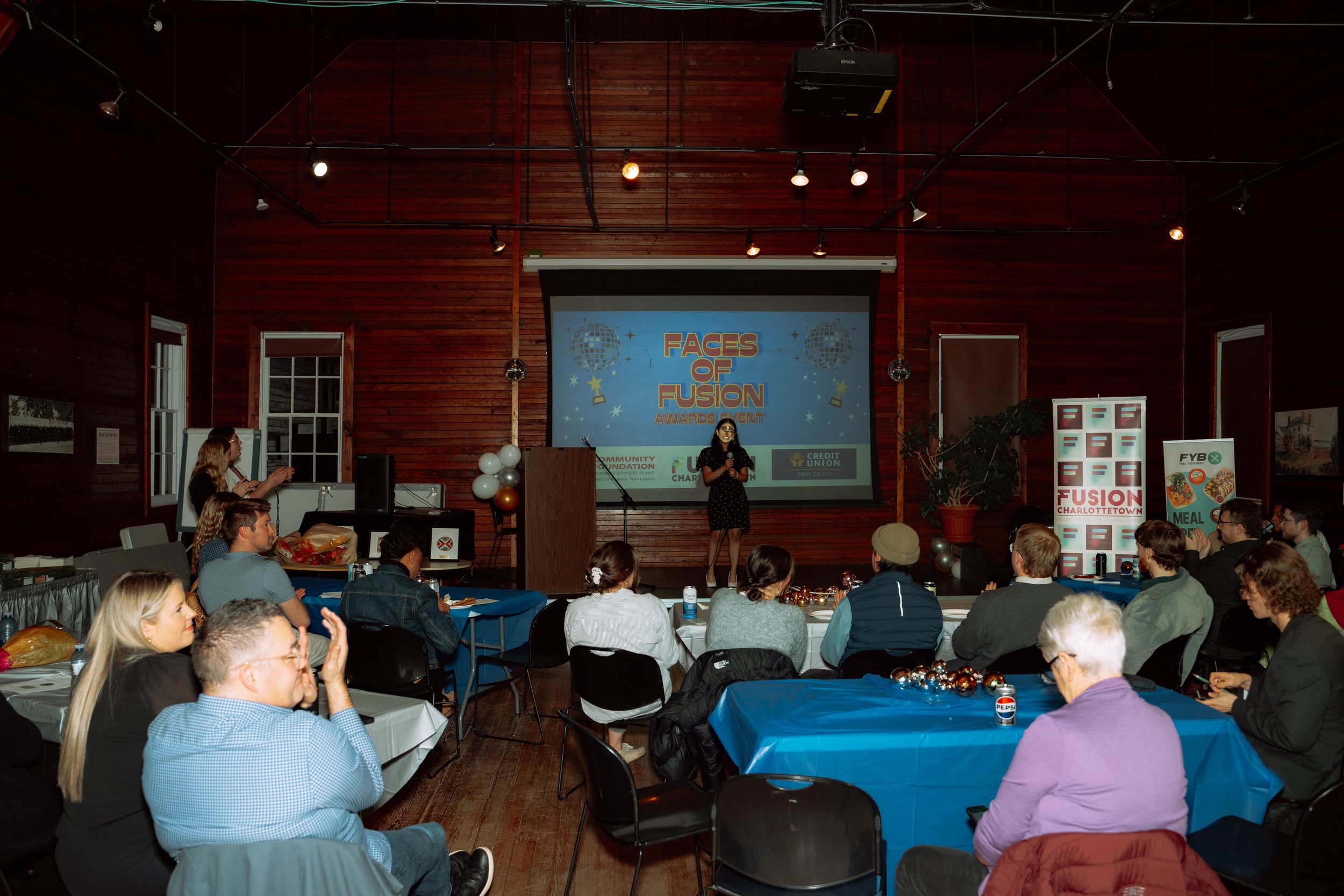 A woman is speaking on a stage at an awards event titled 'Faces of Fusion' with an audience seated at tables in a wood-paneled room. There are decorations and banners related to Fusion Charlottetown.