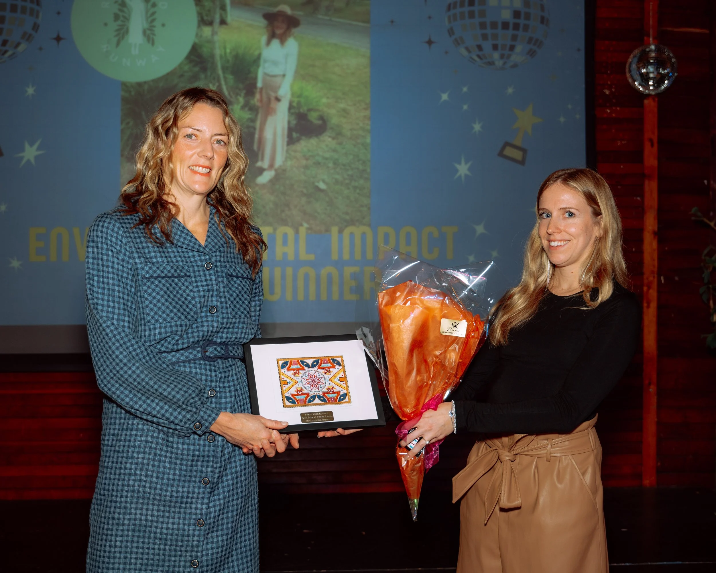 Two women standing on stage, one presenting an award plaque and the other holding a large wrapped bouquet of flowers, during an award ceremony with a screen in the background.