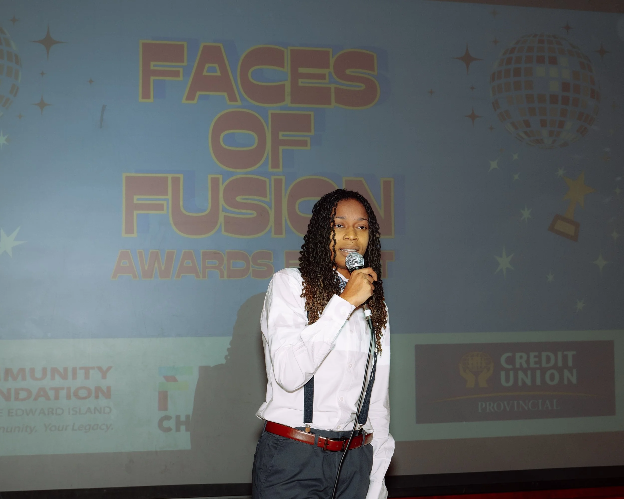A woman in a white shirt and dark pants holding a microphone on stage during an award event titled "Faces of Fusion."