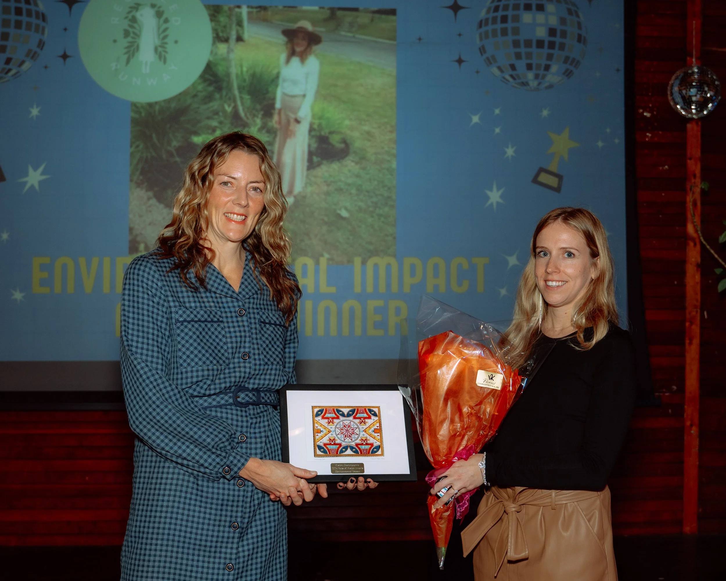 Two women standing in front of a presentation screen, one holding a framed award and the other holding a bouquet of flowers. The woman on the left wears a blue checkered dress, and the woman on the right wears a black top and tan pants.