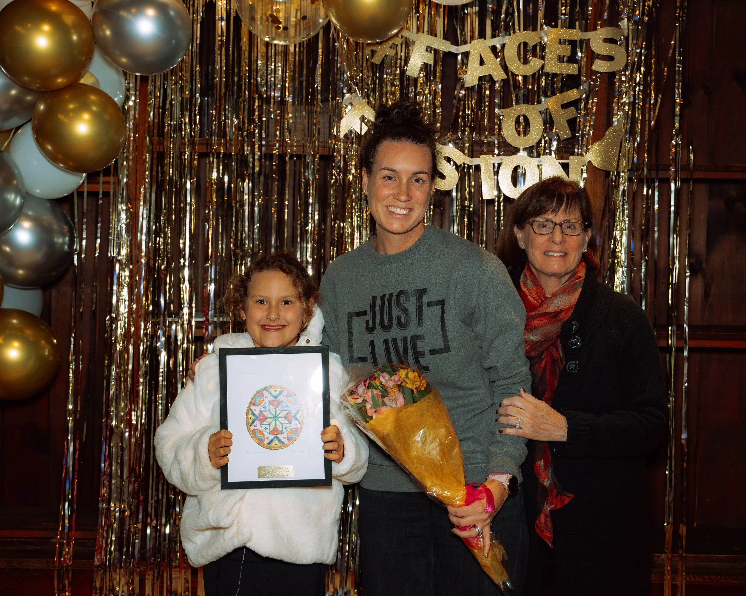 A woman, a young girl, and an older woman pose together in front of a festive backdrop with gold and white balloons and metallic streamers. The girl holds a framed artwork, the woman in the middle has flowers, and all are smiling.