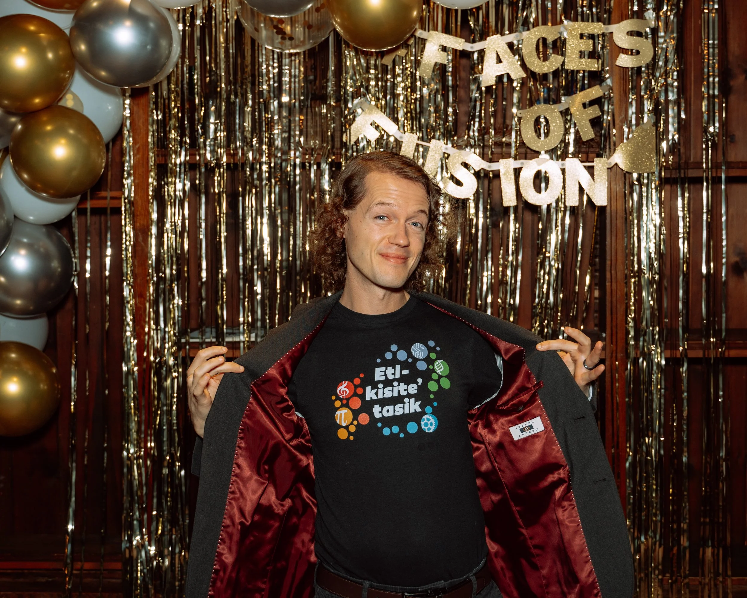 A man with curly hair, wearing a black T-shirt with colorful graphics and text, is standing in front of a shiny gold and silver balloon backdrop with a banner that says "Faces of Houston". He is pulling open his jacket to reveal the T-shirt.