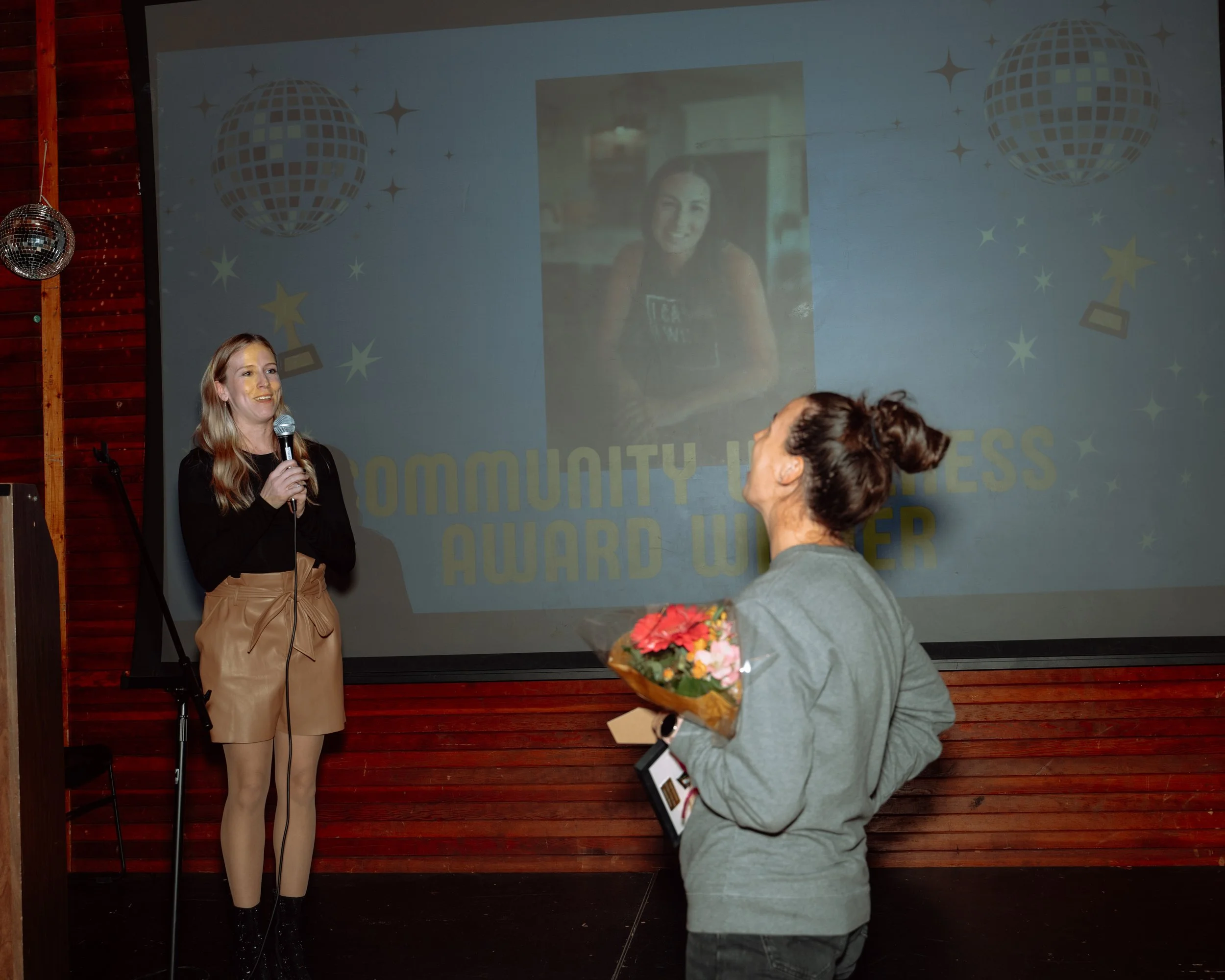 Woman with long blonde hair holding a microphone and speaking at an award ceremony, woman with dark hair in a bun receiving a bouquet of flowers and an award on stage, large screen behind displaying a woman with dark hair and the words "Community Wel
