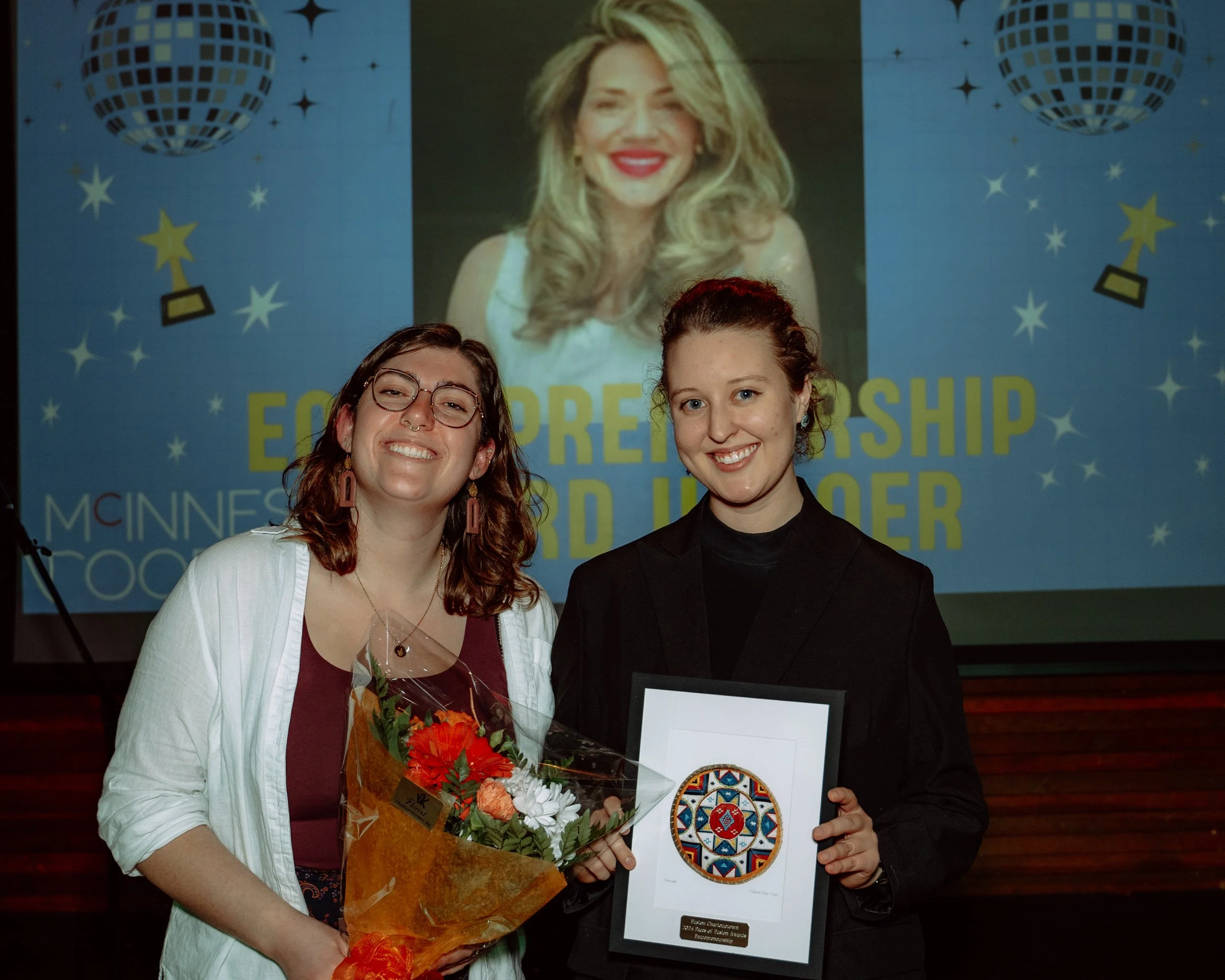 Two women standing together, smiling and holding award and a bouquet of flowers, at an award ceremony with a projected background showing a woman's photo and text about leadership and mentorship.