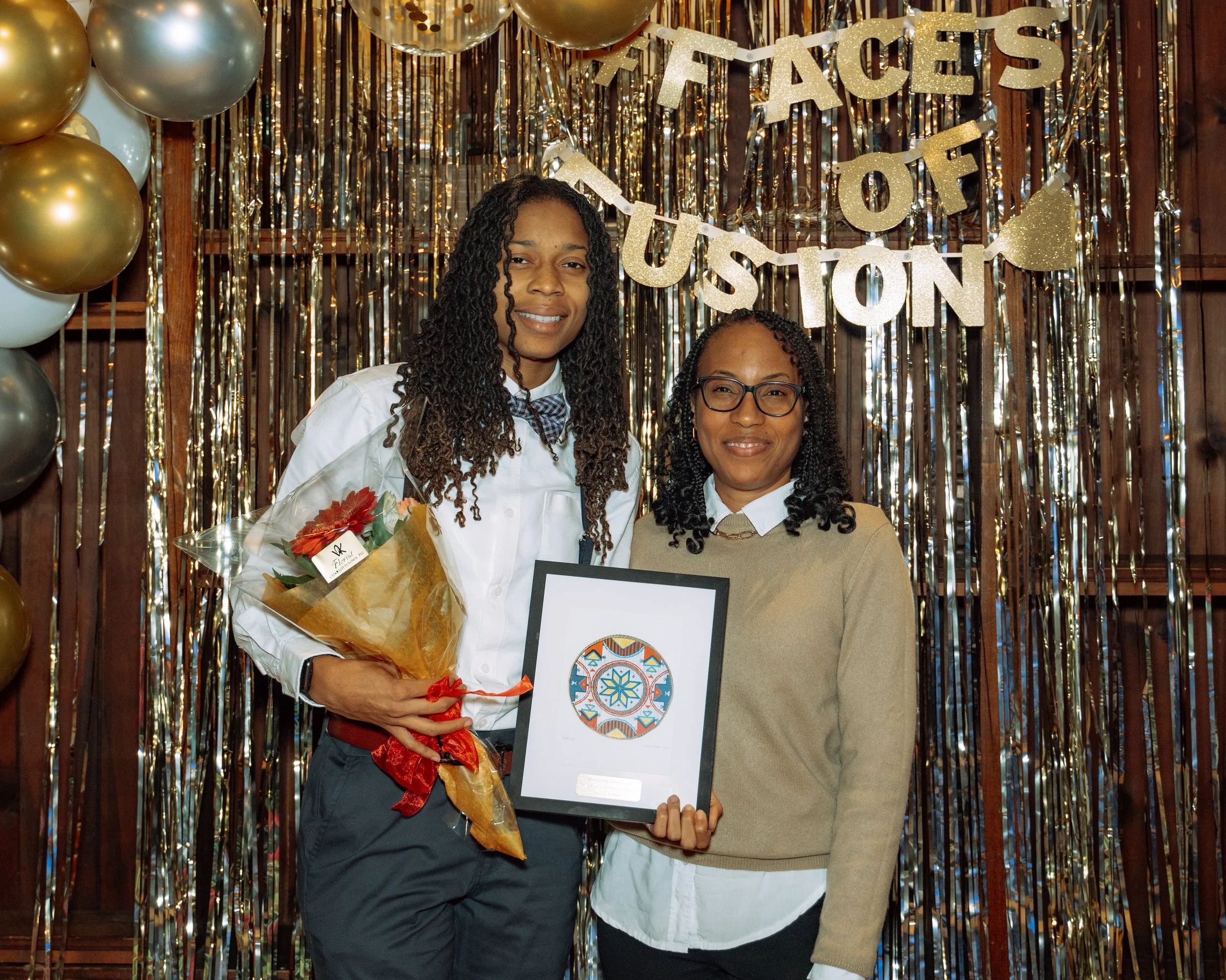 Two women standing together at an event with a golden balloon backdrop and a banner that reads 'Faces of Fusion.' One woman is holding a framed artwork and a bouquet of flowers, and both are smiling.