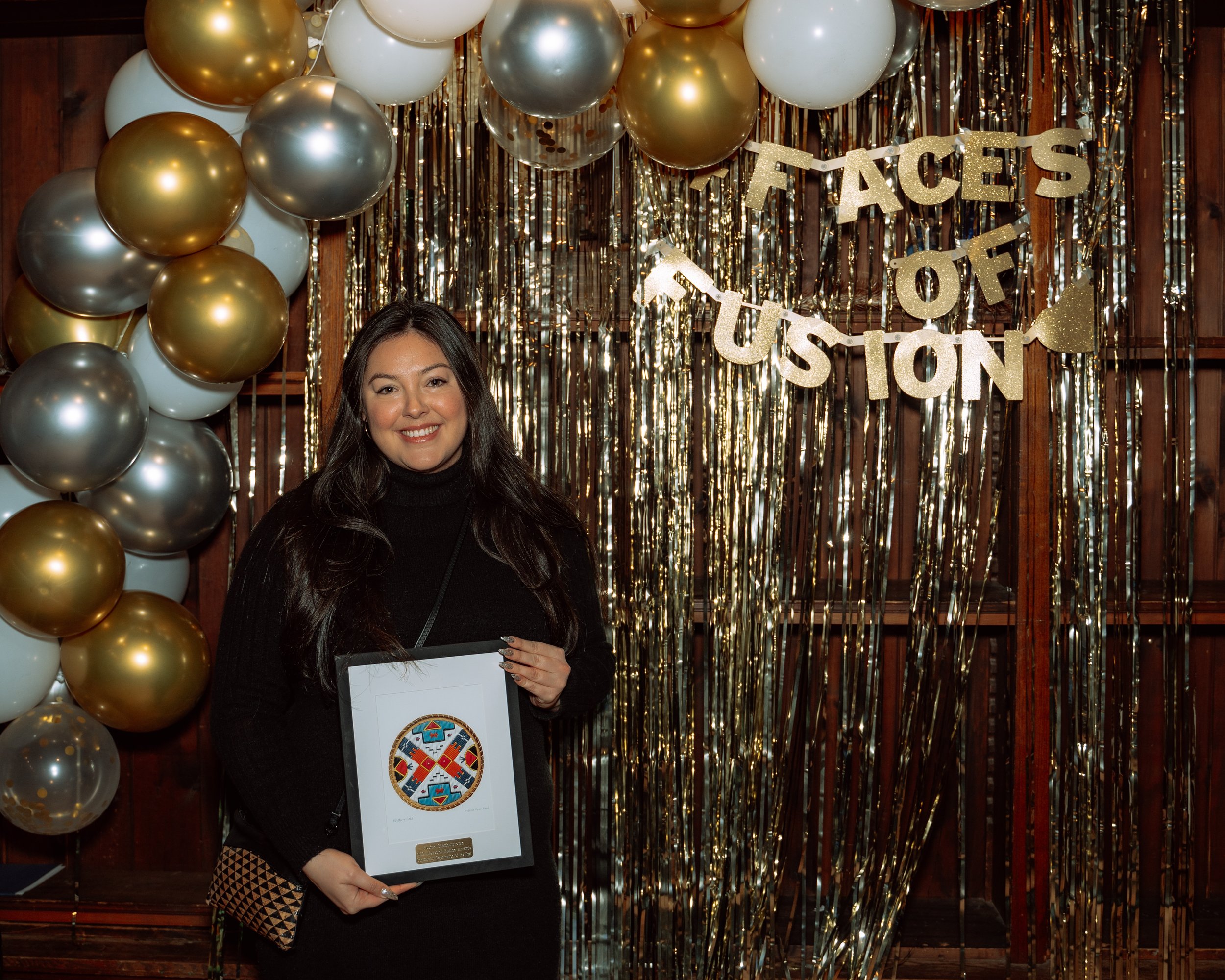 A woman with long dark hair smiling while holding a framed award in front of a gold and silver balloon arch and a gold fringe backdrop. A glittery sign reads 'Faces of Fusion' hanging above her.