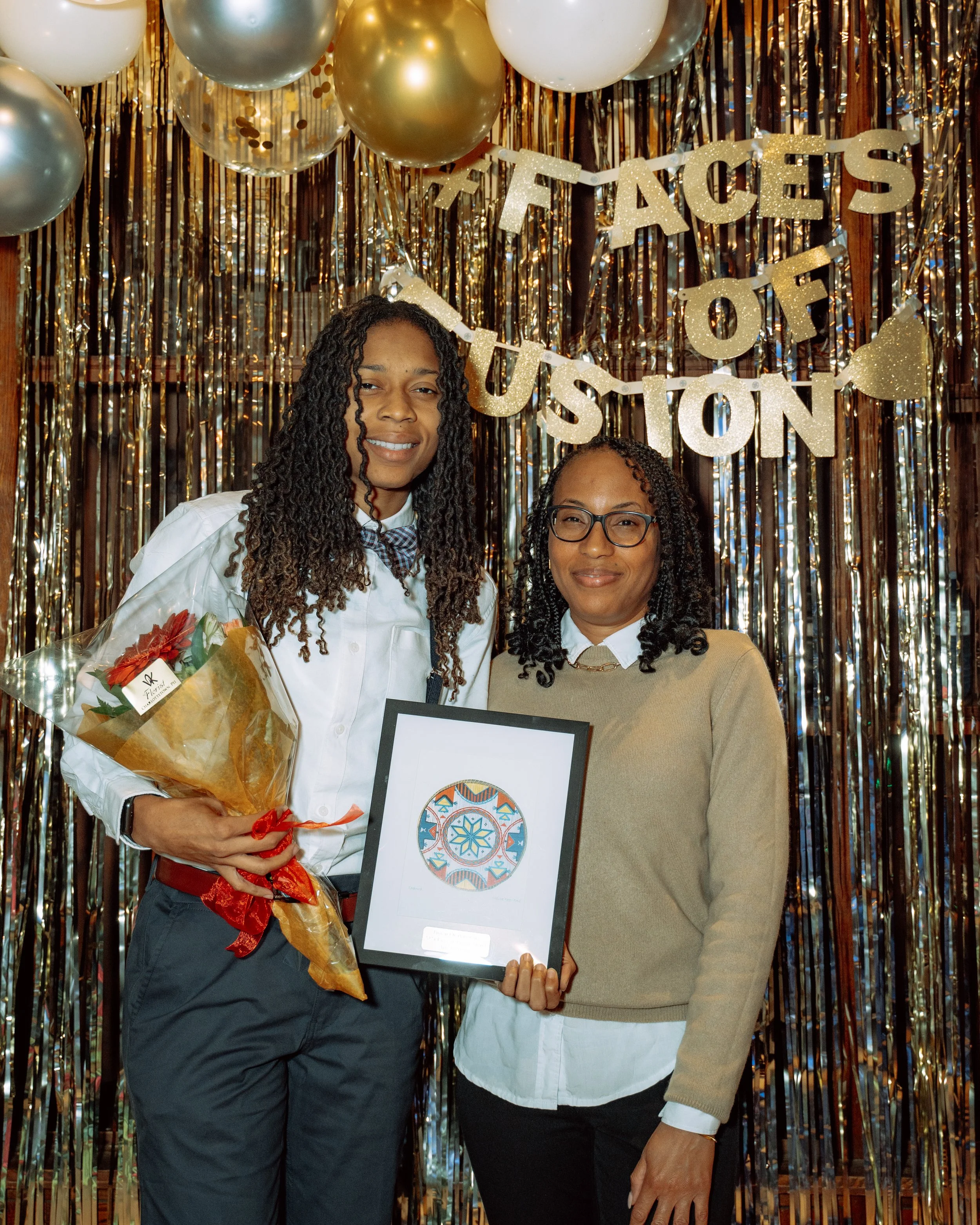 Two women standing in front of a shiny gold and silver backdrop with a balloon decoration and a banner that reads 'Faces of Fusion'. The woman on the left holds a bouquet of flowers and a framed artwork, while the woman on the right holds an empty fr