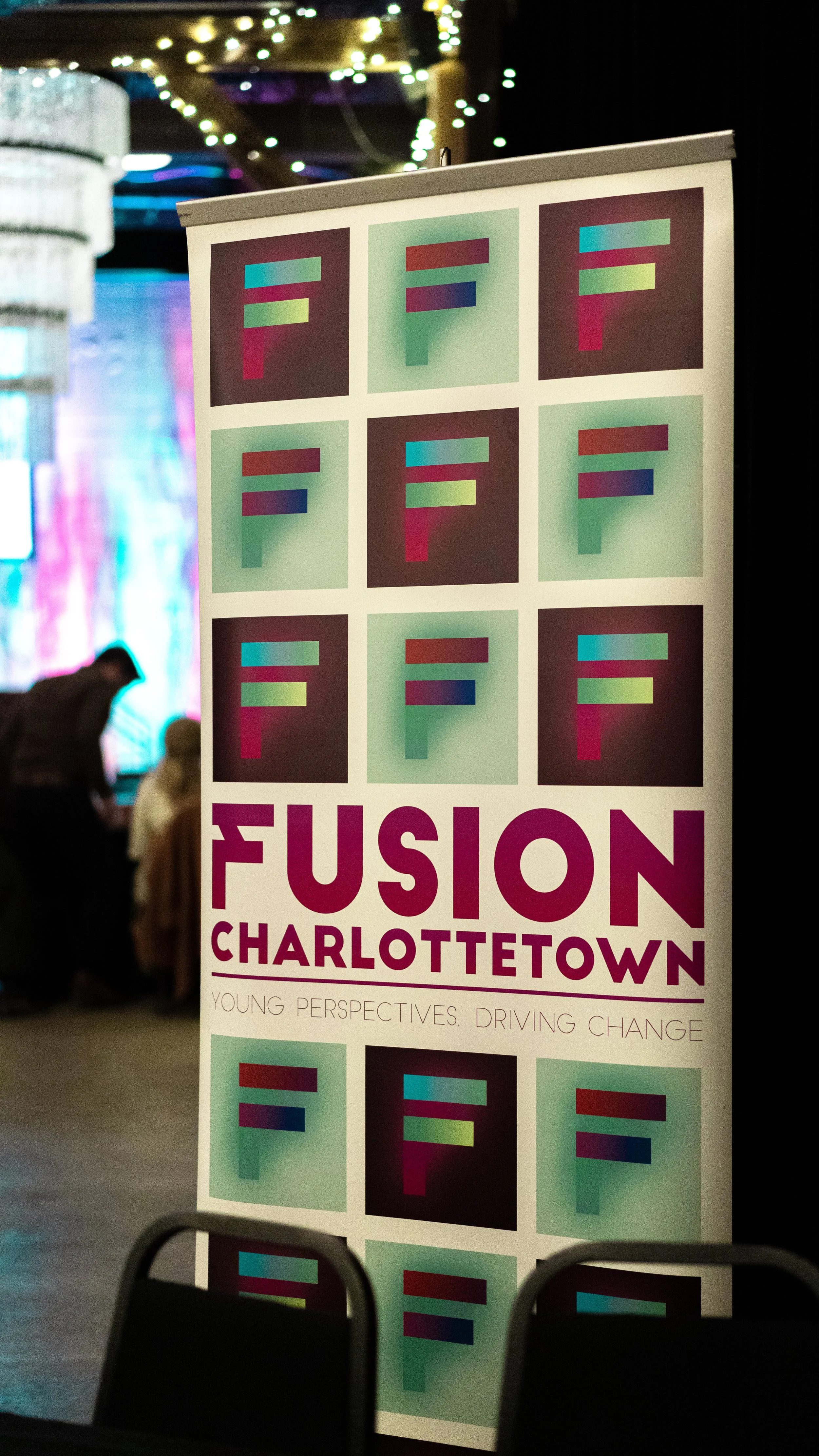 A vertical banner with colorful geometric designs and text that reads "FUSION CHARLOTTETOWN" with the tagline "YOUNG PERSPECTIVES. DRIVING CHANGE" in a dimly lit indoor setting, with blurred people and decorative lights in the background.