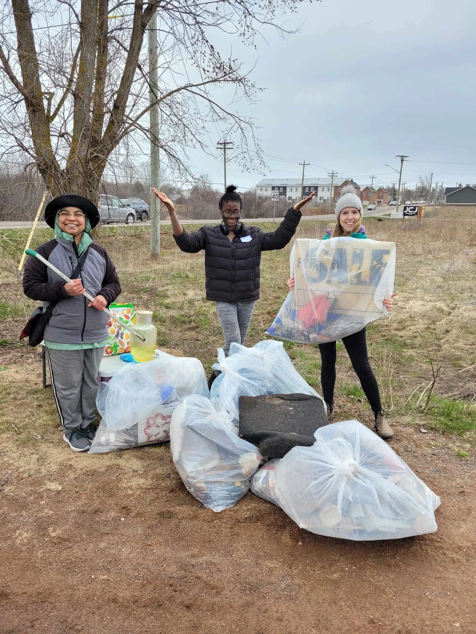 Three women participating in a community cleanup outdoors, standing behind trash bags filled with collected litter, with a tree and a roadside in the background.