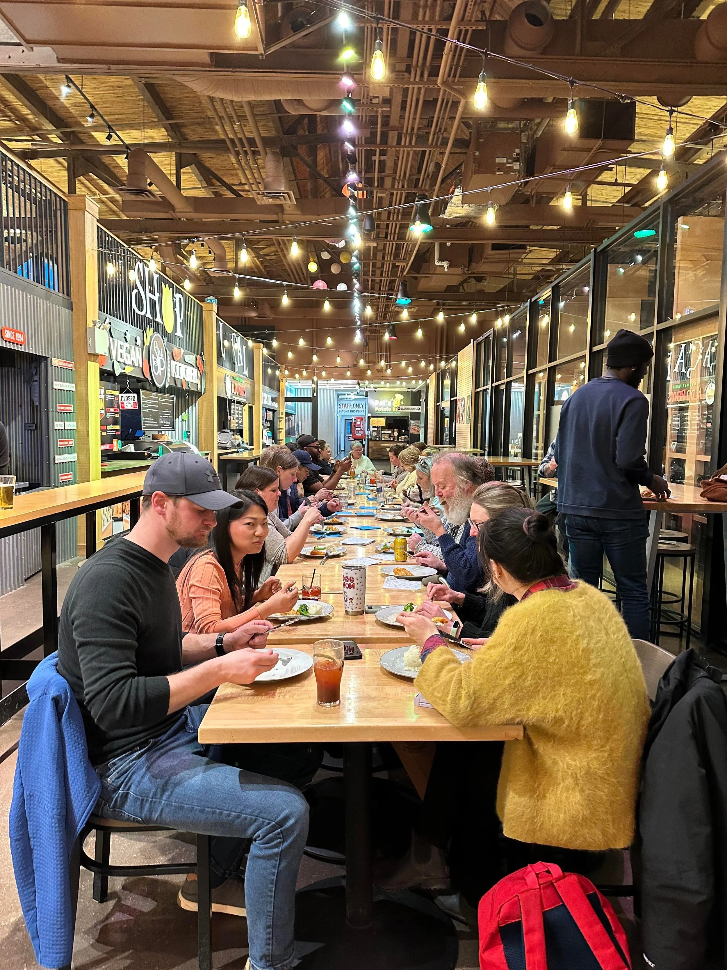People dining at a long wooden table inside a restaurant with string lights hanging from the ceiling, and a counter area with signboards in the background.