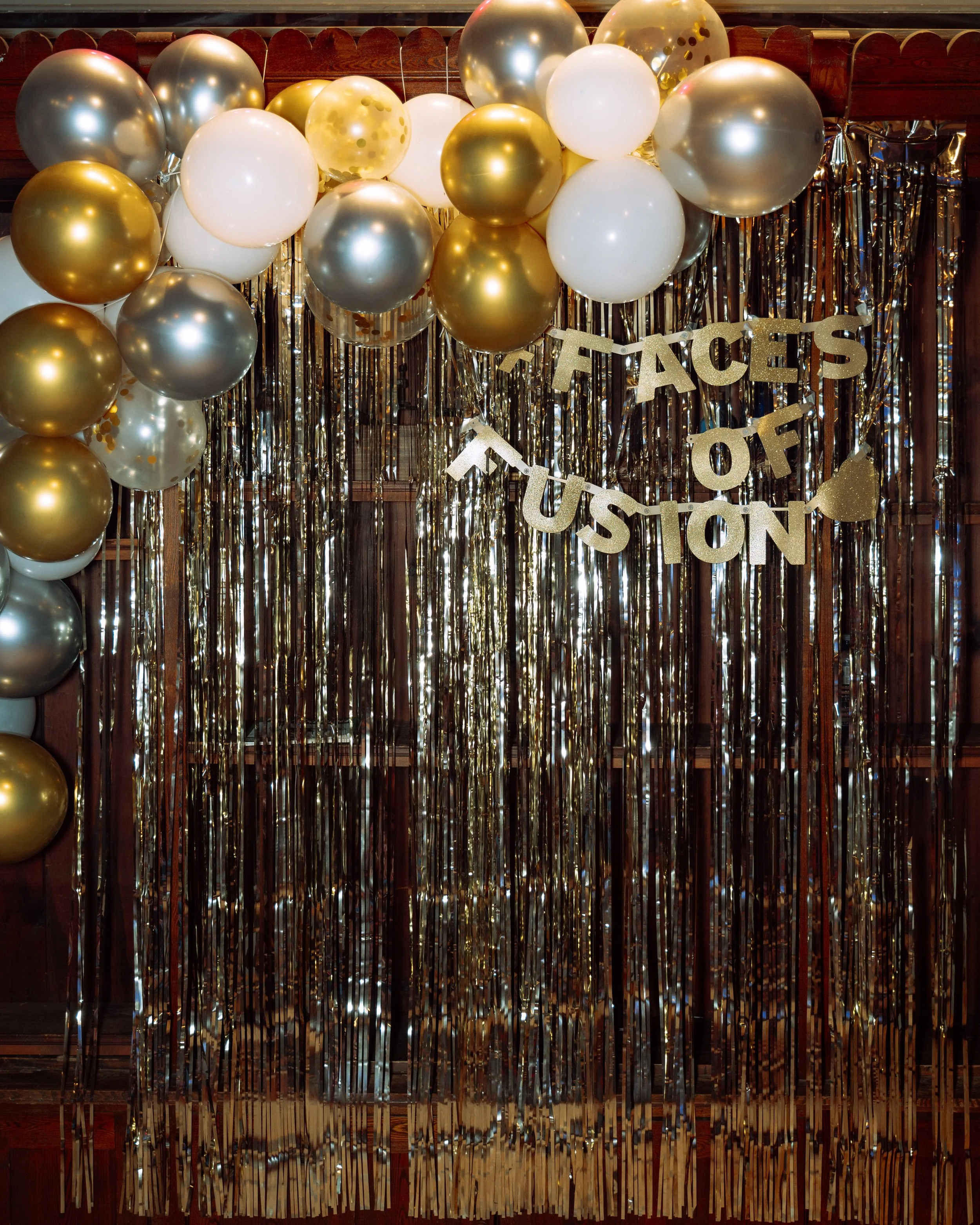 Decorative backdrop with gold, silver, and white balloons, metallic fringe curtains, and a banner that reads "Faces of Tustin" for celebration or event.