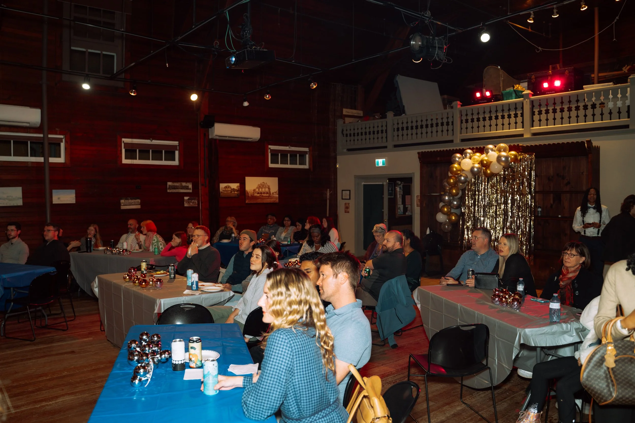 People sitting at decorated tables during a festive indoor event with a balloon and curtain backdrop.