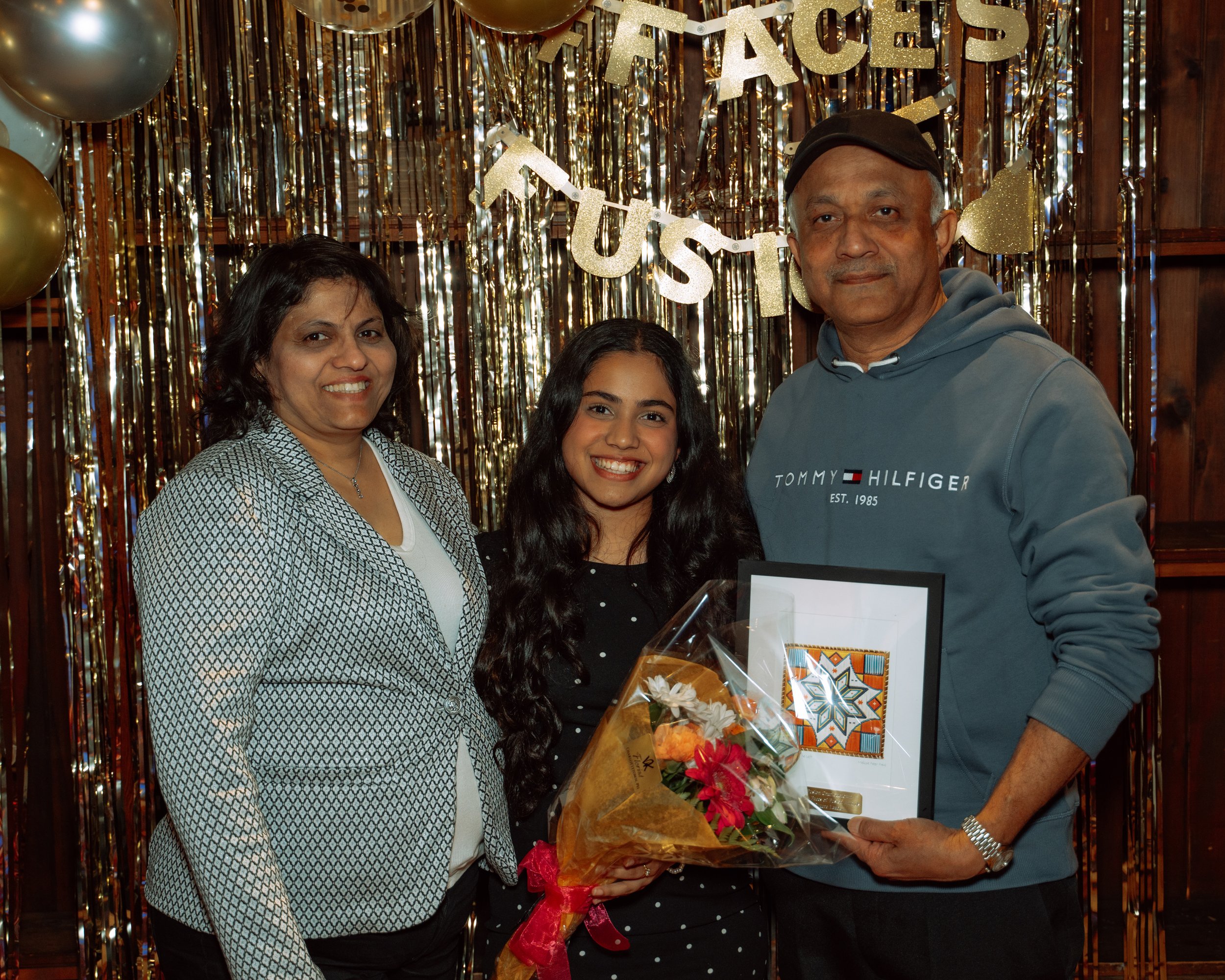 A joyful family celebration with three people standing in front of a gold and silver metallic fringe backdrop. The backdrop has balloons and a banner that reads 'TUES'. The woman in the middle is holding a bouquet of flowers and a framed artwork, and
