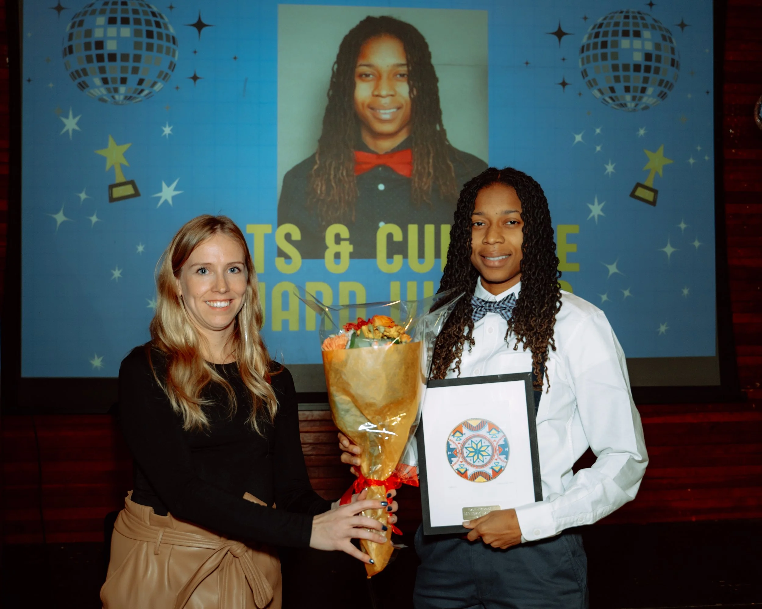 A young woman with long curly hair in a white shirt with a bow tie and dark pants holding a framed certificate, standing next to an older woman with long blonde hair in a black top and beige skirt, holding a bouquet of flowers. They are at an award c