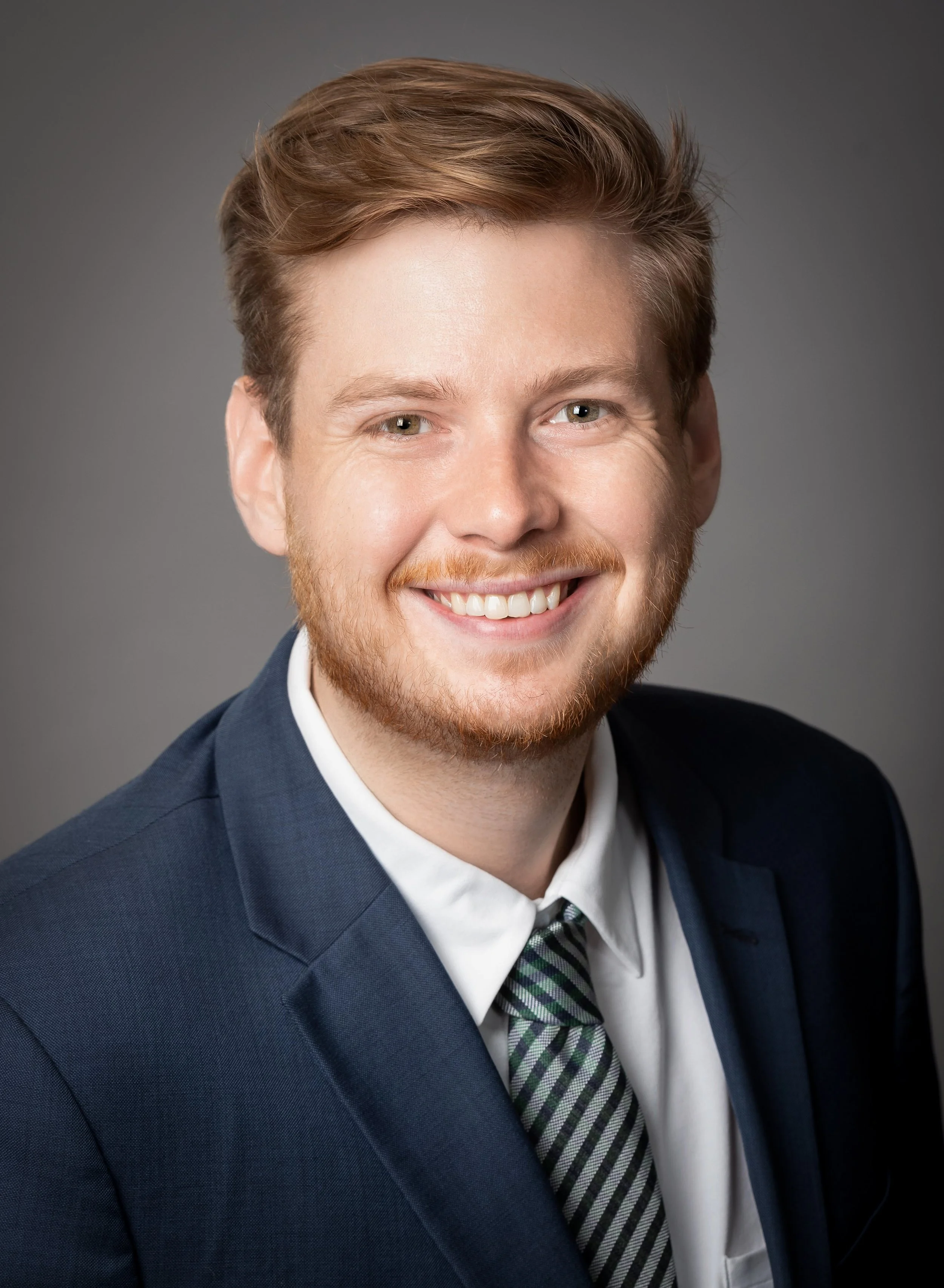 A smiling young man with reddish hair and a beard, wearing a navy blue suit, white shirt, and a gray striped tie, posed against a gray background.