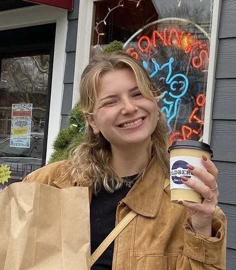 A smiling blonde woman standing in front of a store holding a cup of coffee