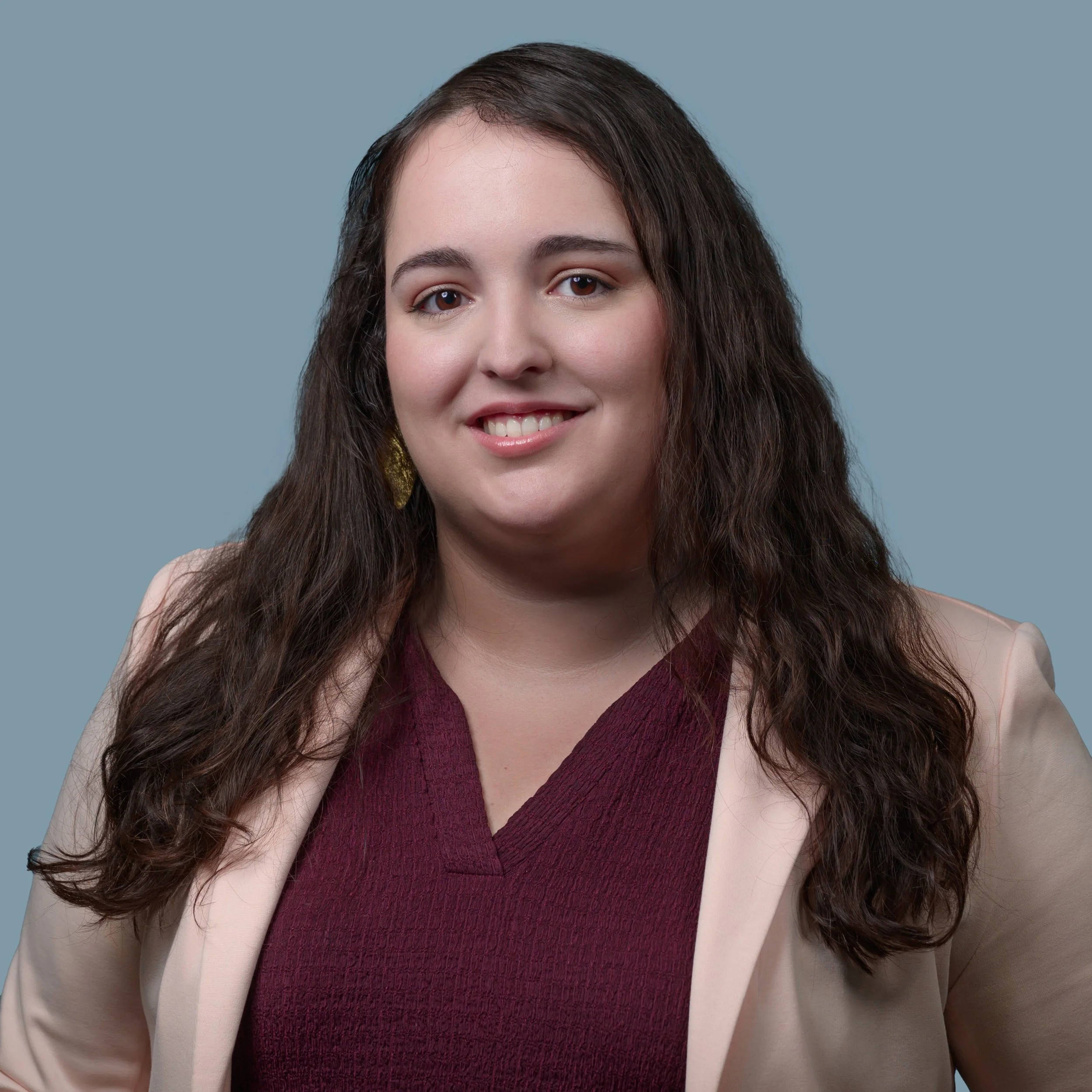 A woman with medium brown hair wearing a pink blazer and burgundy blouse against a gray background.
