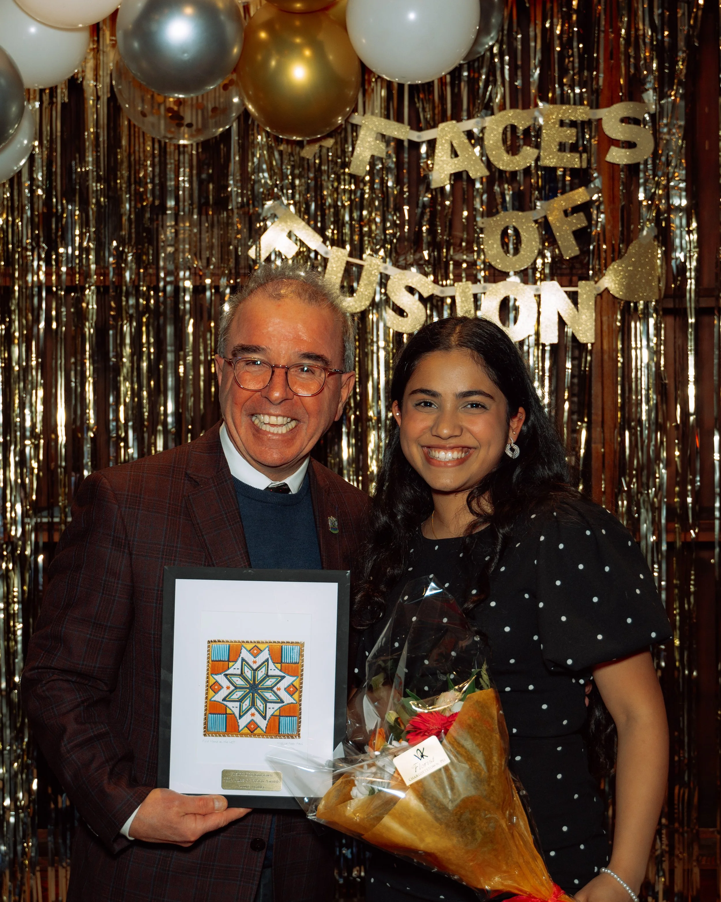 A man and a woman smiling at a celebration with a decorative backdrop that says 'Faces of Fusion.' The man is holding a framed piece of artwork, and the woman is holding a bouquet of flowers.