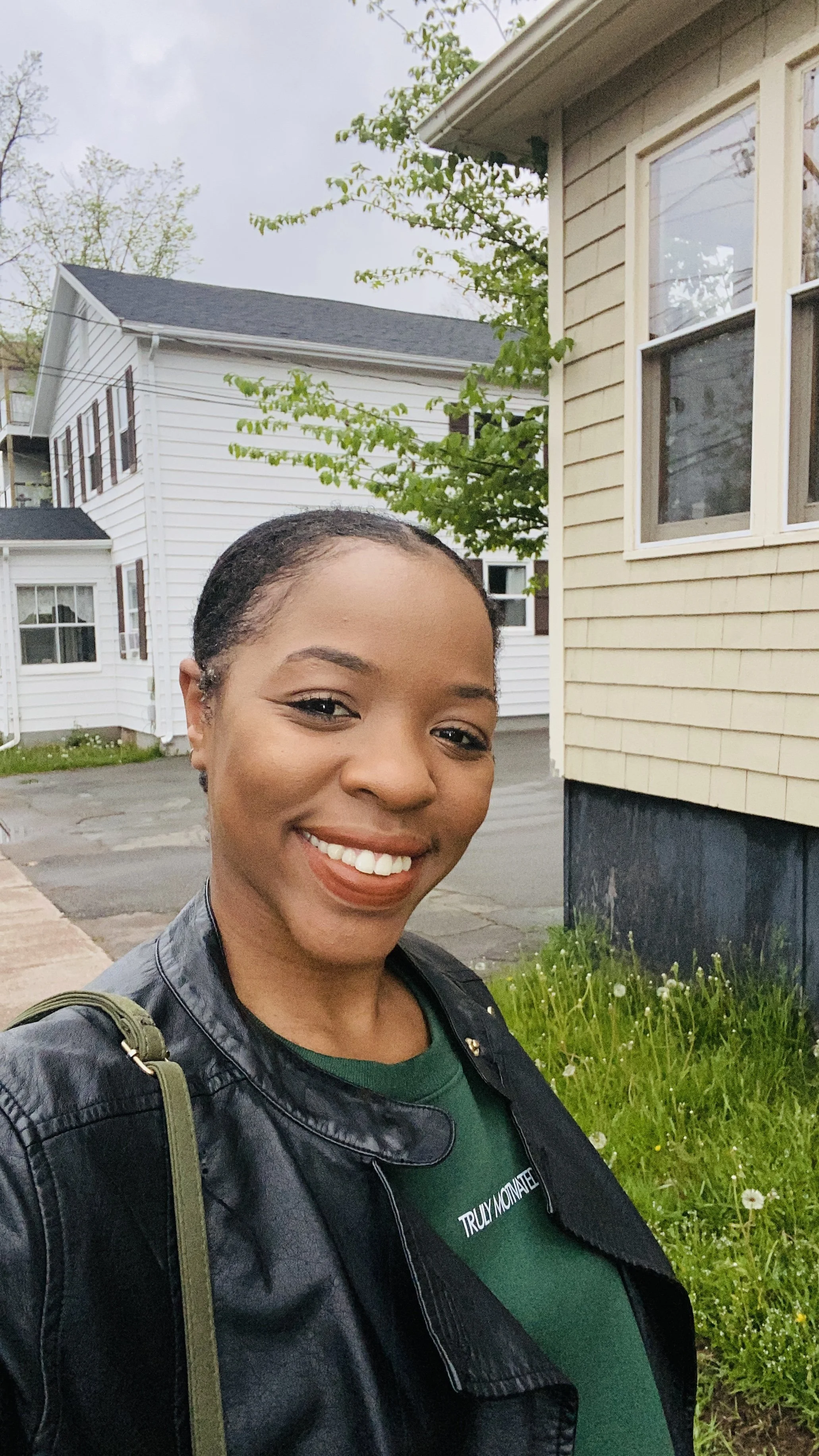A woman smiling in front of a house with siding and windows, on a cloudy day with trees in the background.