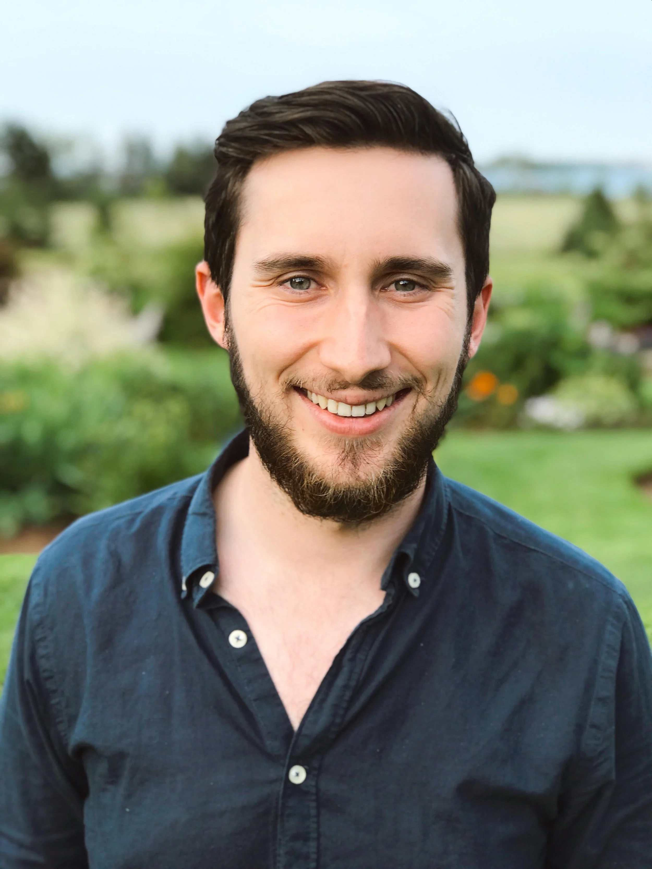 A smiling young man with dark hair and a beard outdoors in a green park or garden.