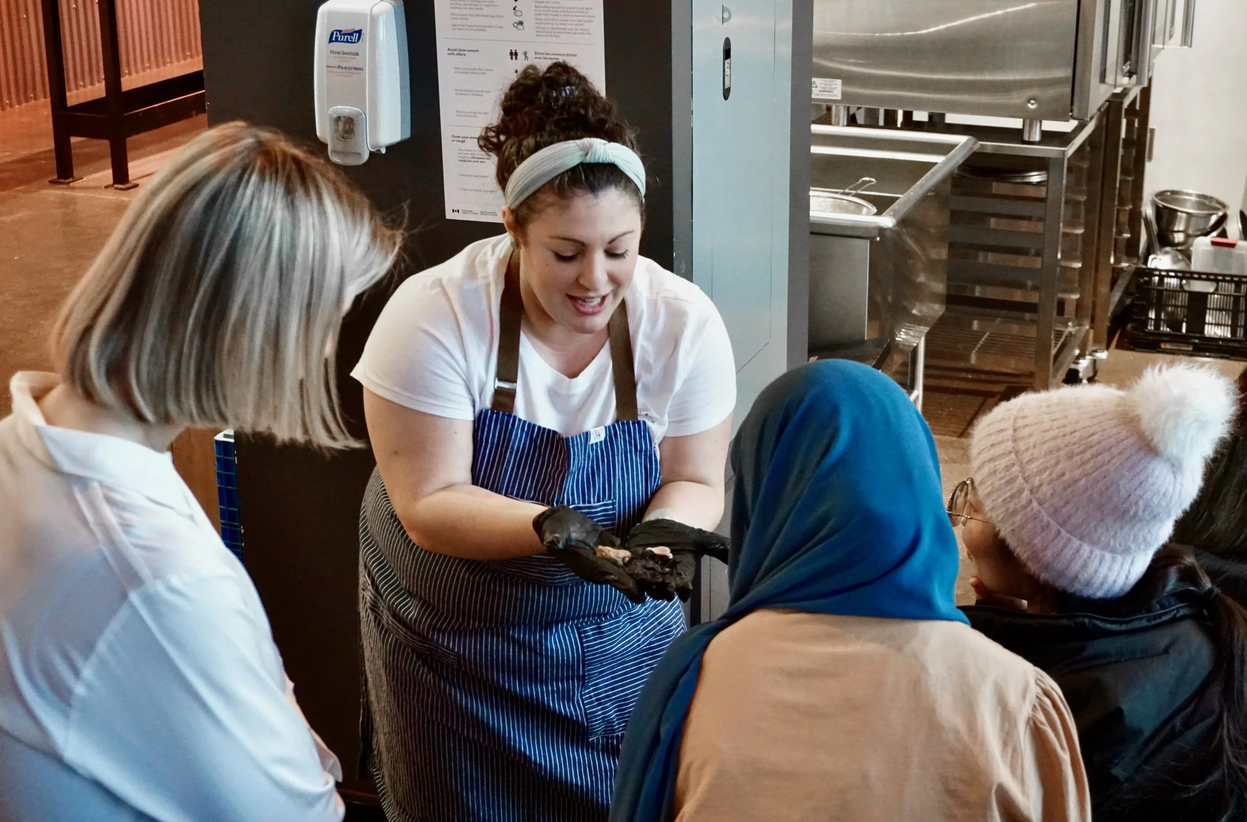 A woman explaining something to a group of women seated around a table in a kitchen or restaurant setting.