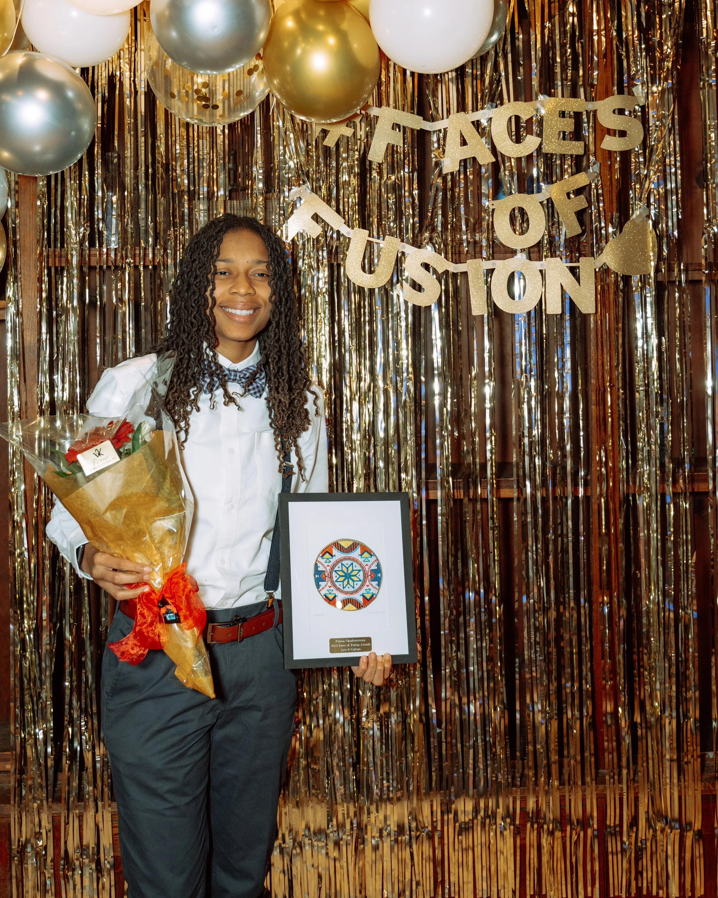 A young girl in a white shirt and gray pants holding a bouquet of flowers and a framed artwork, standing in front of a festive gold and white balloon backdrop with a banner that says 'Faces of Justice'.
