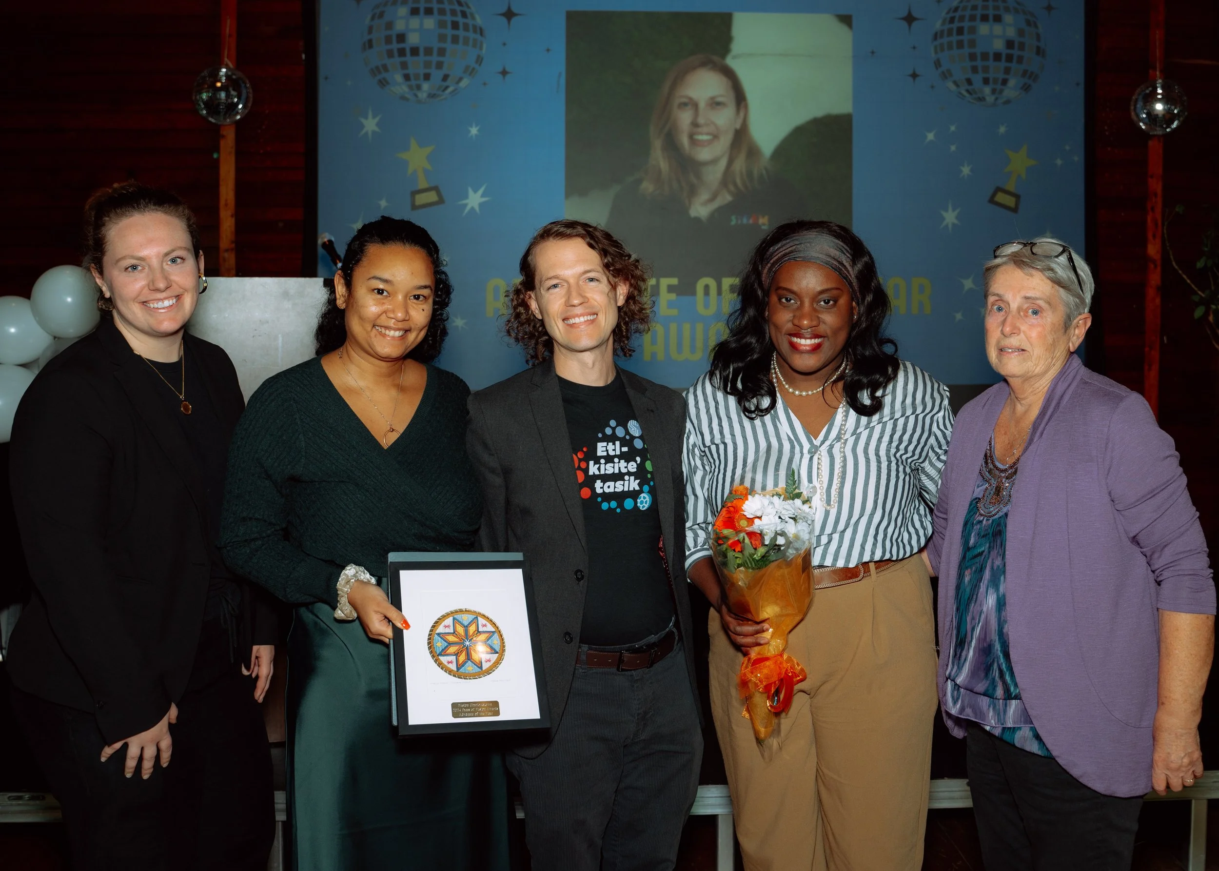 Six women standing together indoors at an award event, one holding a framed certificate and another holding a bouquet of flowers, with a large banner and decorated background behind them.