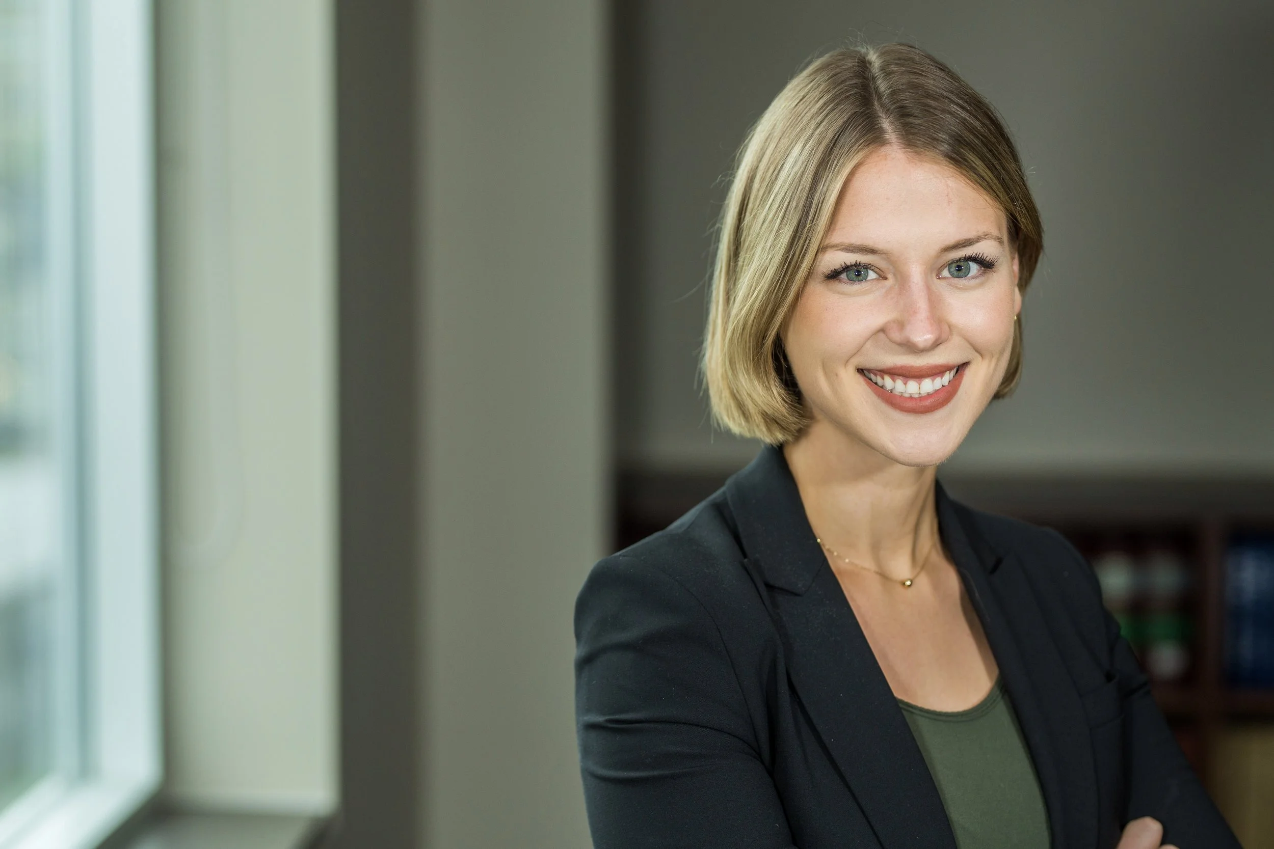 A smiling woman with short blonde hair, wearing a black blazer and a dark green top, standing indoors near a window.