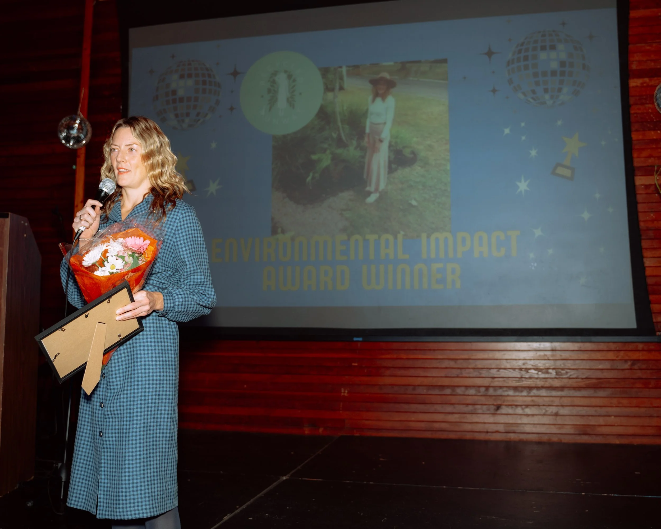 A woman with curly blonde hair, wearing a blue checkered dress, holding a microphone, a bouquet of flowers, and a framed award, speaking at an event. Behind her, a large screen displays an environmental impact award with a photo of her outdoors, near