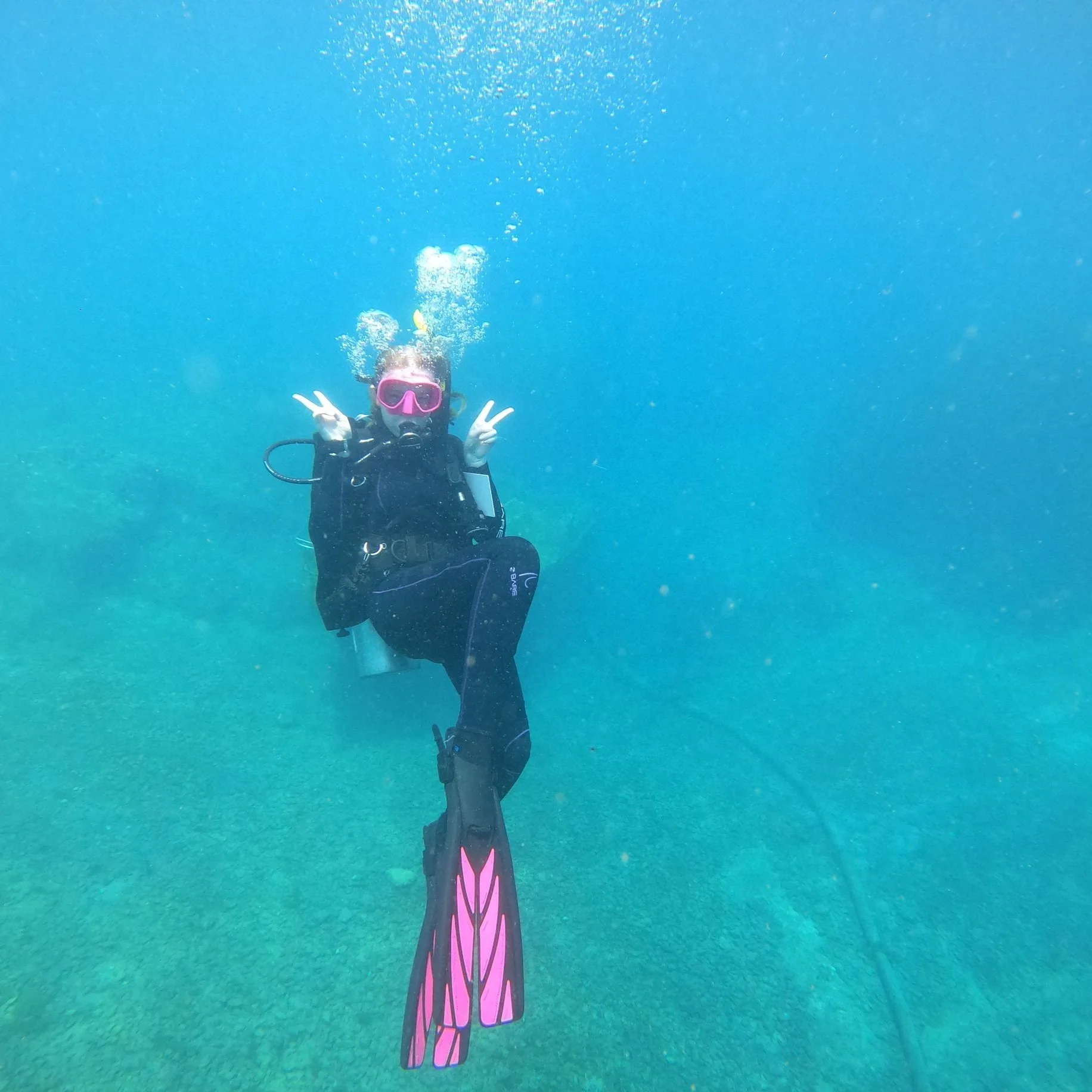 Person scuba diving underwater, wearing a pink mask and fins, making peace signs with both hands.