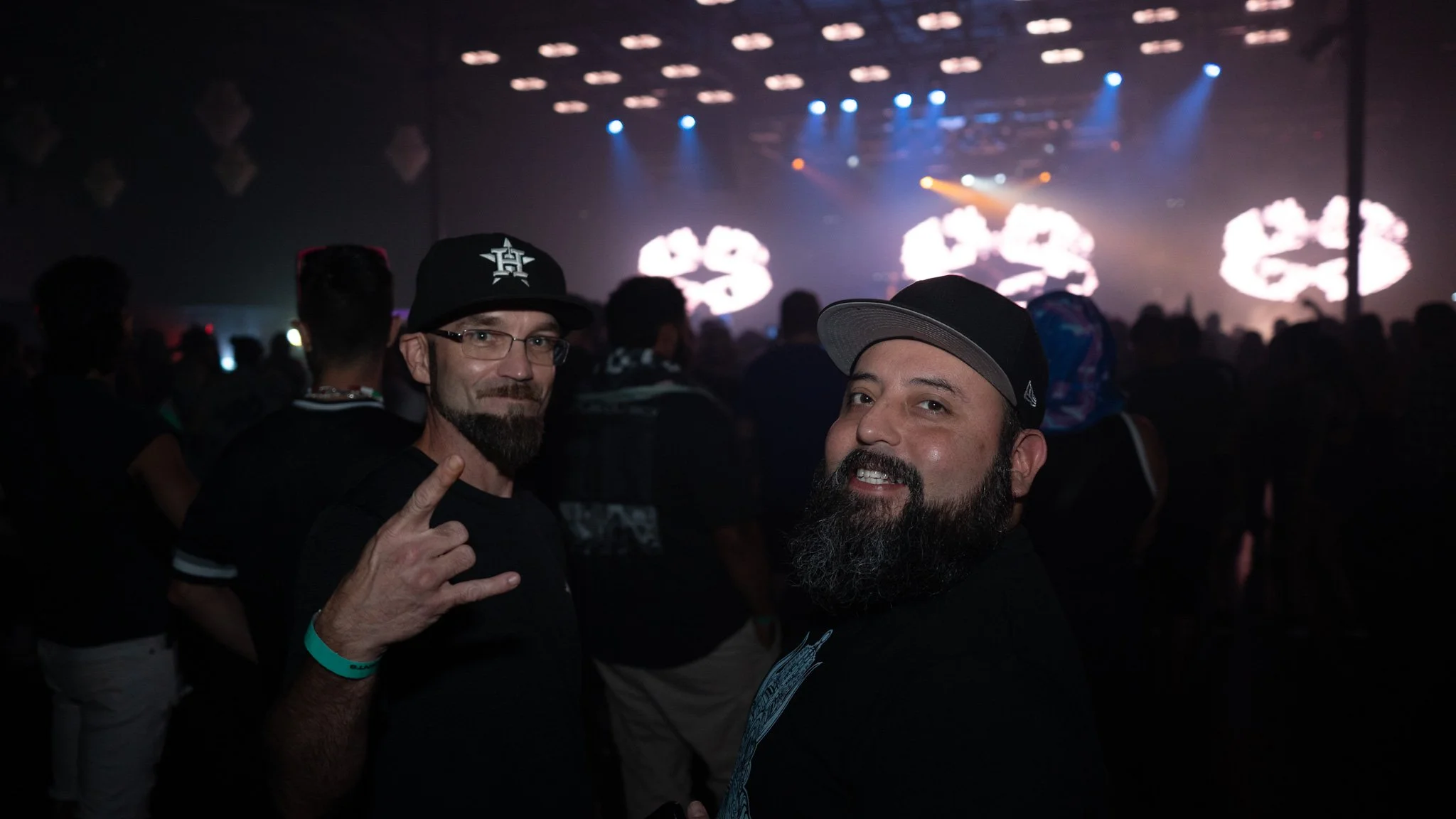 Two men with beards and black clothing at a concert or music festival, with a dark background and stage lights.