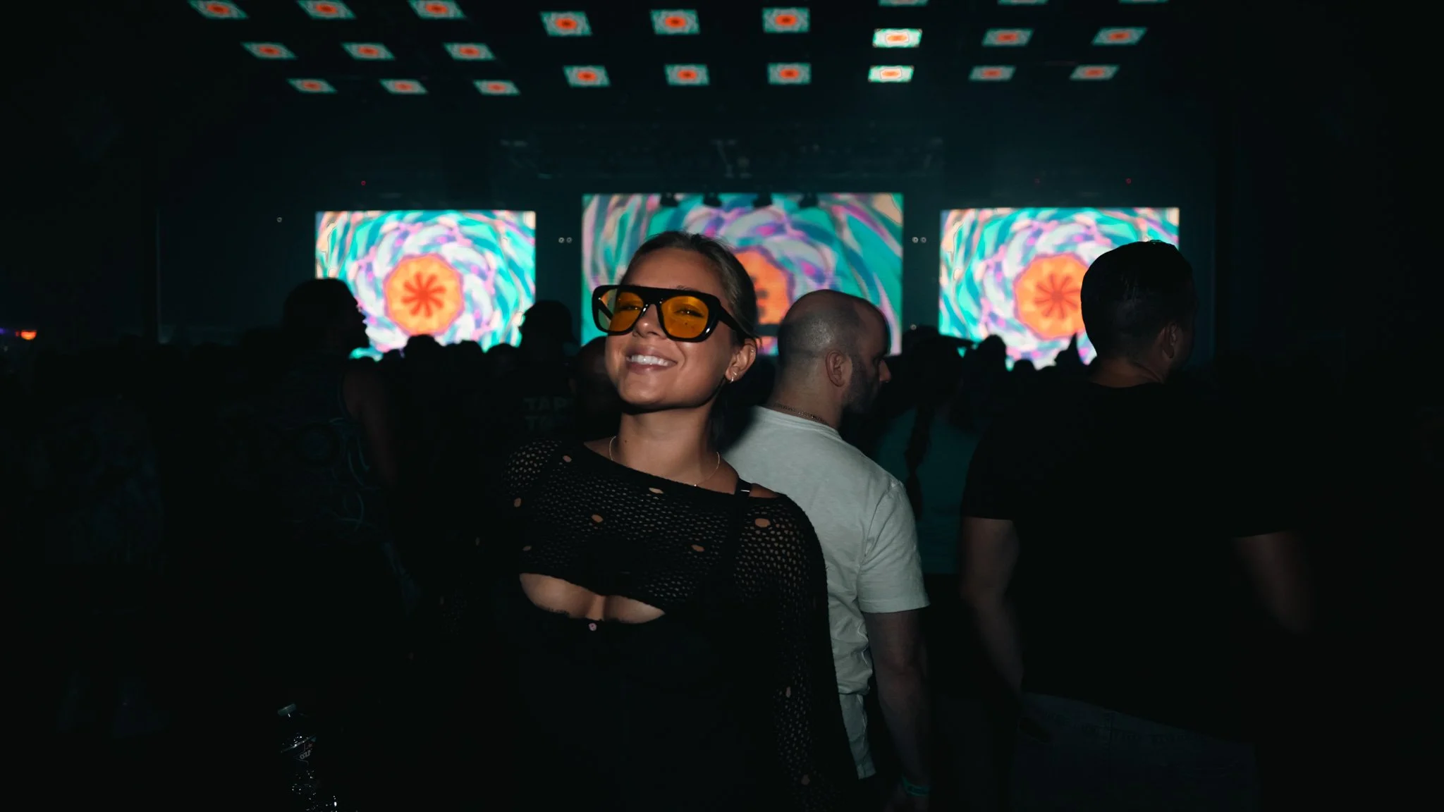 A woman smiling and wearing tinted glasses at a crowded indoor concert or electronic music event, with bright abstract visuals on large screens in the background.