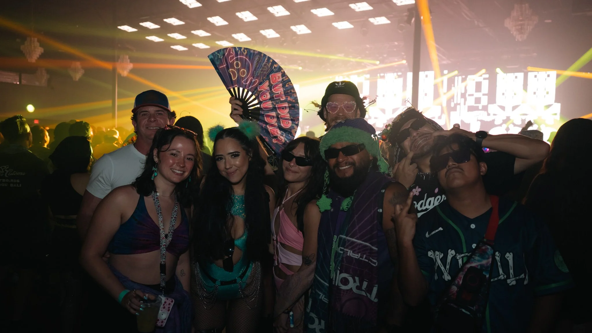 Group of people at a concert or festival, posing for a photo with colorful stage lighting in the background, many wearing sunglasses and casual or festival attire.