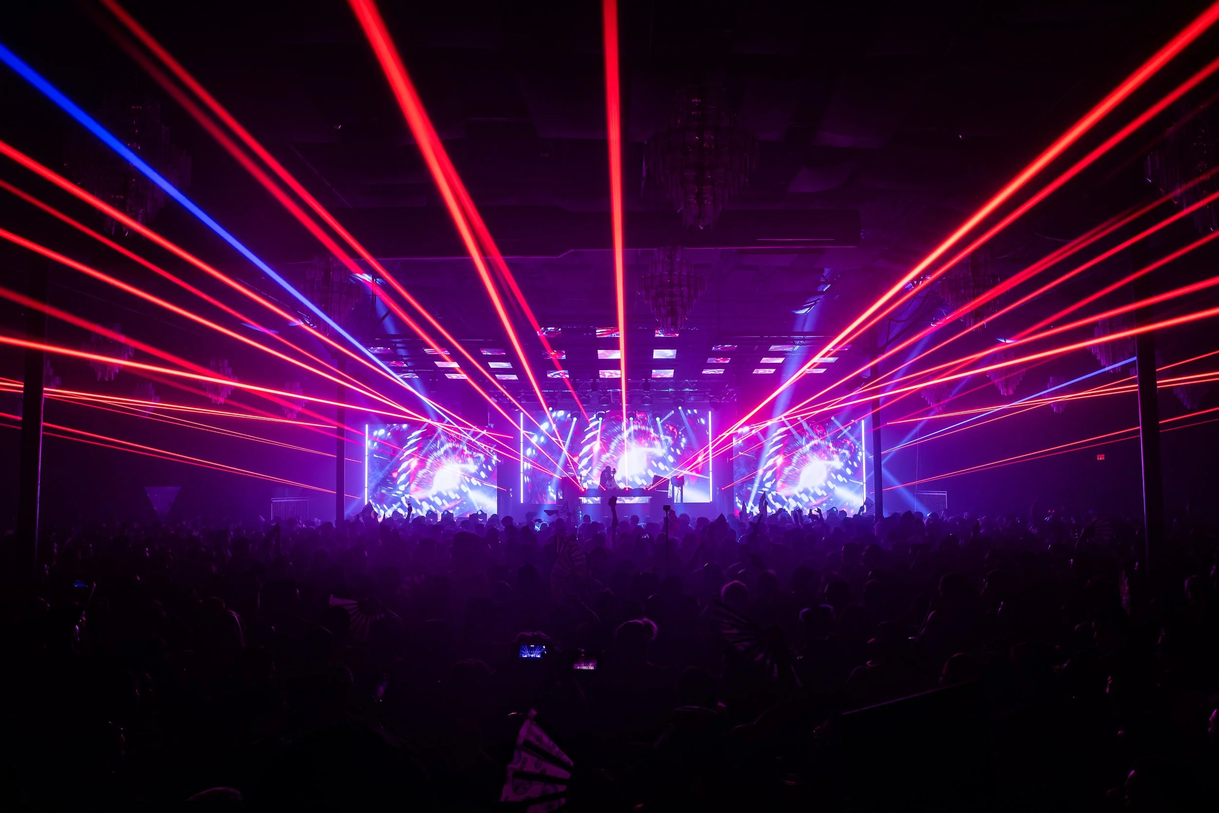 A concert or nightclub scene with colorful laser lights and a large crowd, with stage screens displaying dynamic visuals.