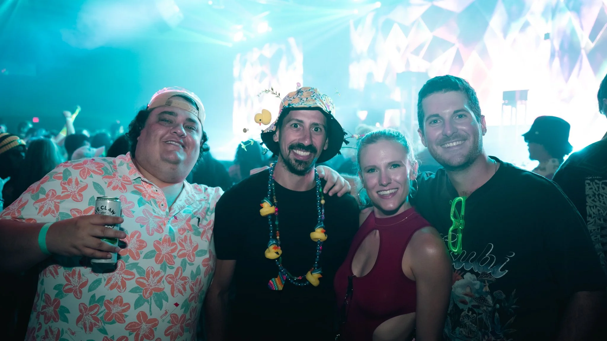 Group of four people at a concert or festival enjoying nightlife, with colorful lights and a crowded background.