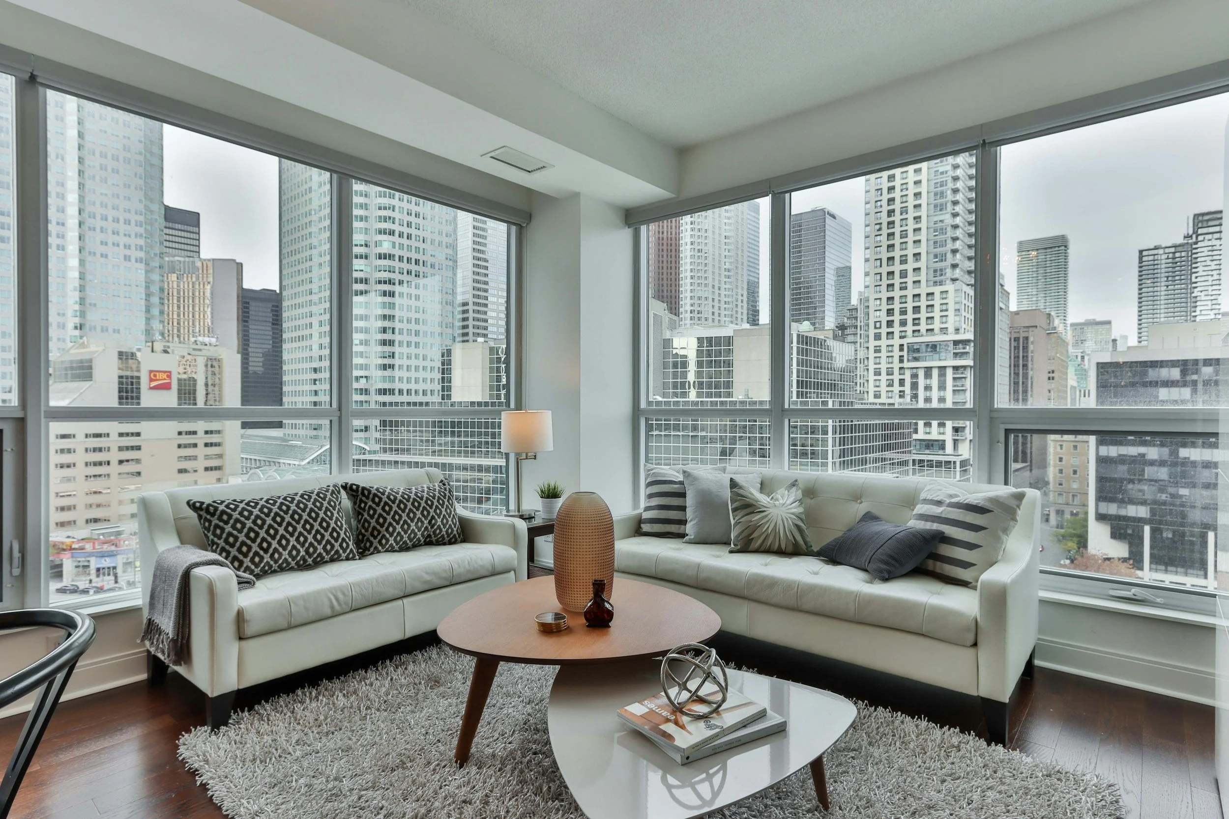 Living room with large windows showing city skyline, two white sofas with patterned pillows, a coffee table with decorative items, a side table with a lamp, and a shaggy area rug.