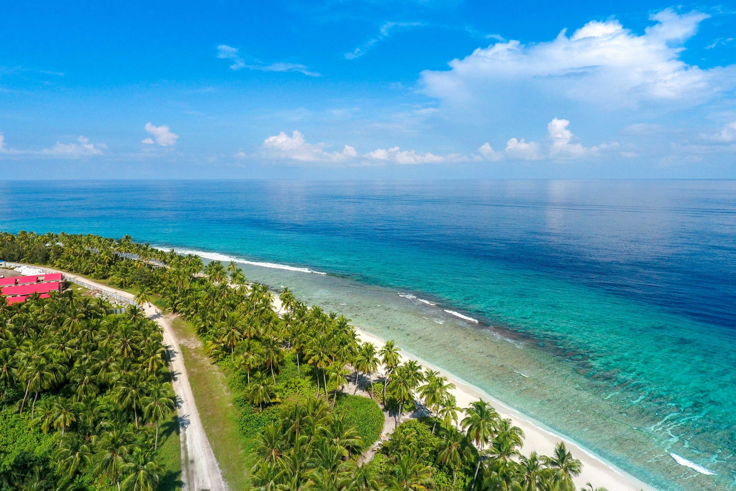 Aerial view of a tropical beach with white sand, lush green palm trees, and clear turquoise water, under a blue sky with some clouds.