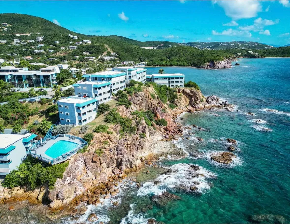 Aerial view of modern white and blue buildings perched on rocky cliffs along a clear blue ocean coastline with green hills in the background.