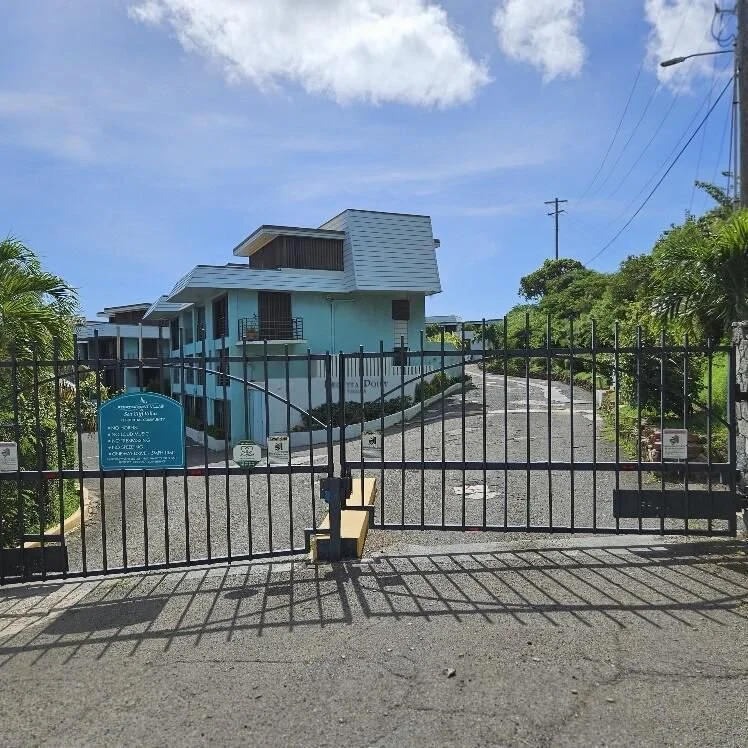 Gated entrance to a residential complex with a blue building in the background, surrounded by greenery and a clear sky.