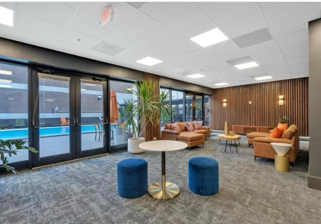 Indoor lounge area with beige sofas, a round marble table, blue poufs, a potted plant, and large windows overlooking a swimming pool.